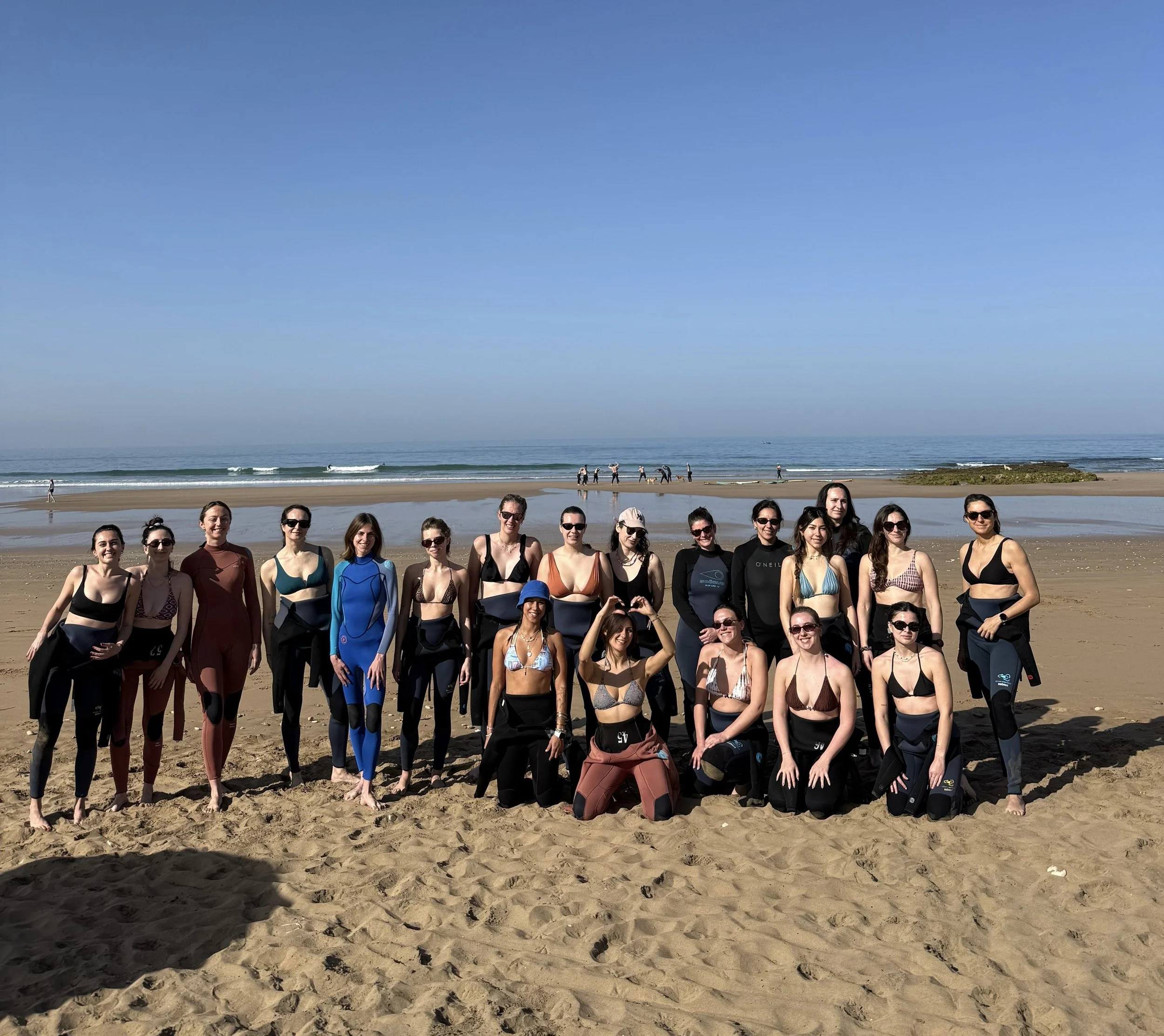 Groupe de femmes sur la plage, assises et debout, portant des combinaisons de plongée et des bikinis, avec la mer et le ciel en arrière-plan.