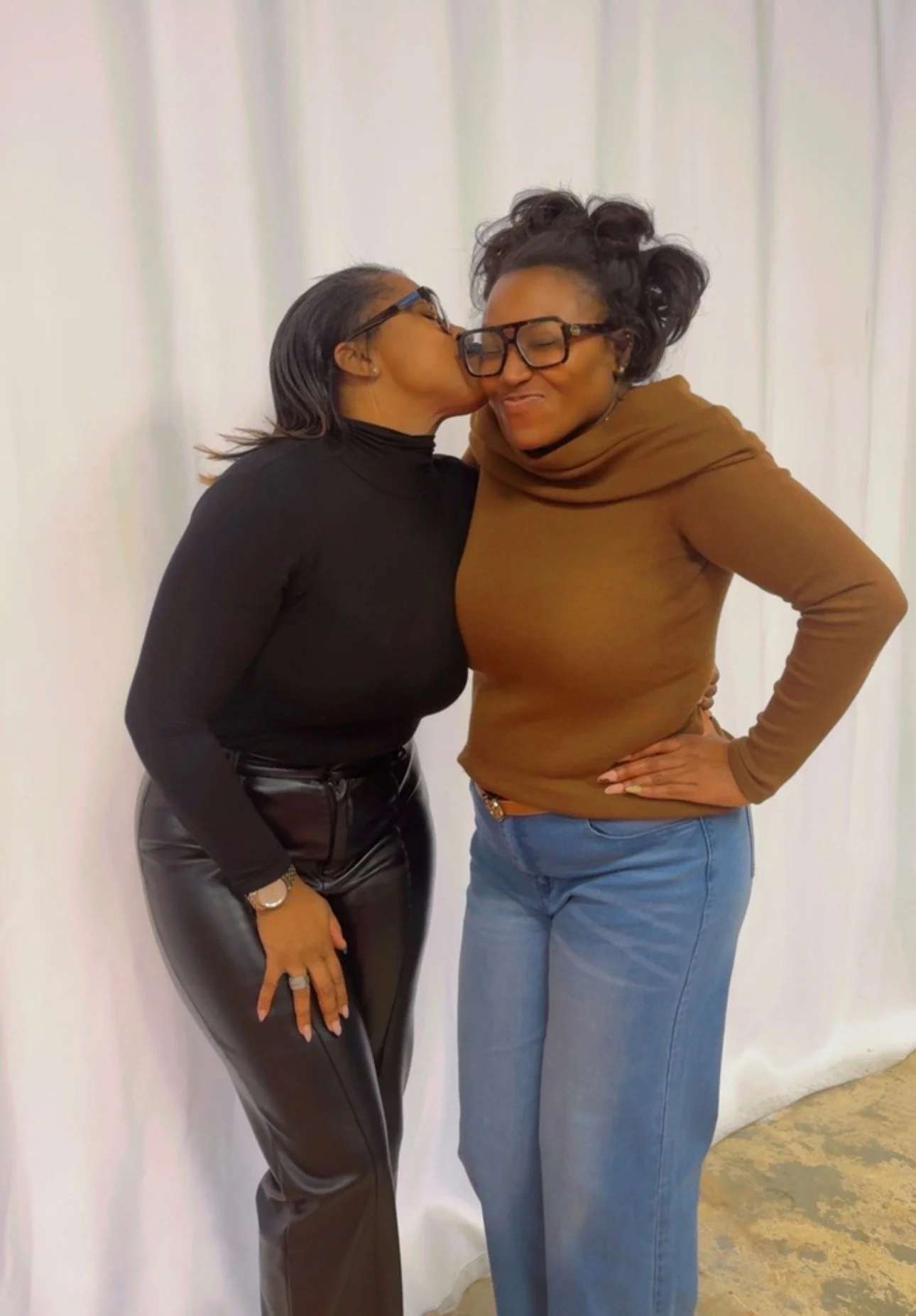 Two women, one on the left wearing a black turtleneck and leather pants, the other on the right wearing glasses, a brown top, and blue jeans, sharing a moment of affection, with the woman on the left giving a kiss on the cheek of the woman on the right.