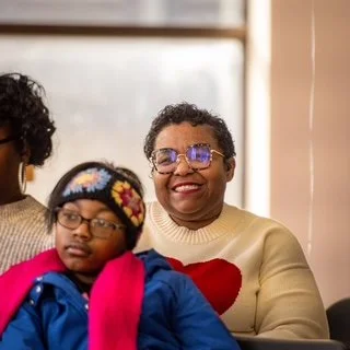 A woman smiling and wearing glasses sits next to a young girl with glasses and a colorful beanie. Both are indoors.