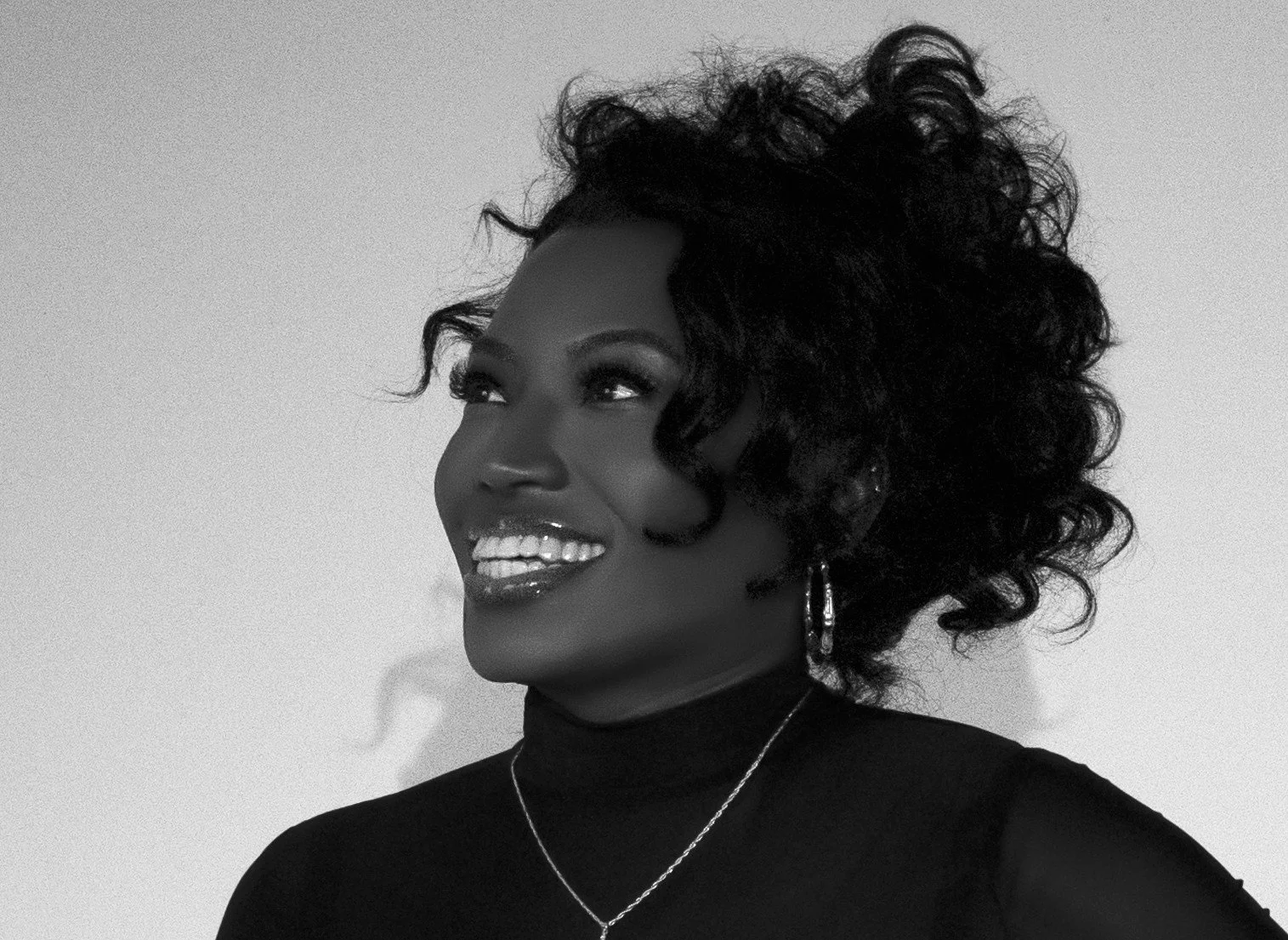 Black and white portrait of a smiling woman with curly hair, wearing a black top, earrings, and a necklace, against a plain background.