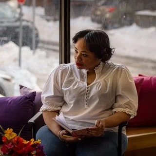 Woman sitting indoors holding papers near window with snowy landscape outside.