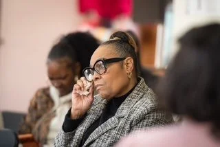 A woman in glasses adjusting her eyeglasses in a classroom setting with other women in the background.