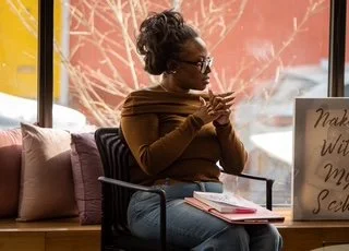 A woman sitting on a couch inside a cafe, looking at her phone with pillows behind her and a window showing a leafless tree outside.