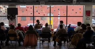 A group of people seated in chairs facing a large window with pink wall behind it, decorated with small heart-shaped stickers or decorations. Some people are taking notes or listening attentively.