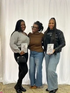 Three women standing together, smiling, holding books, in front of a white curtain.