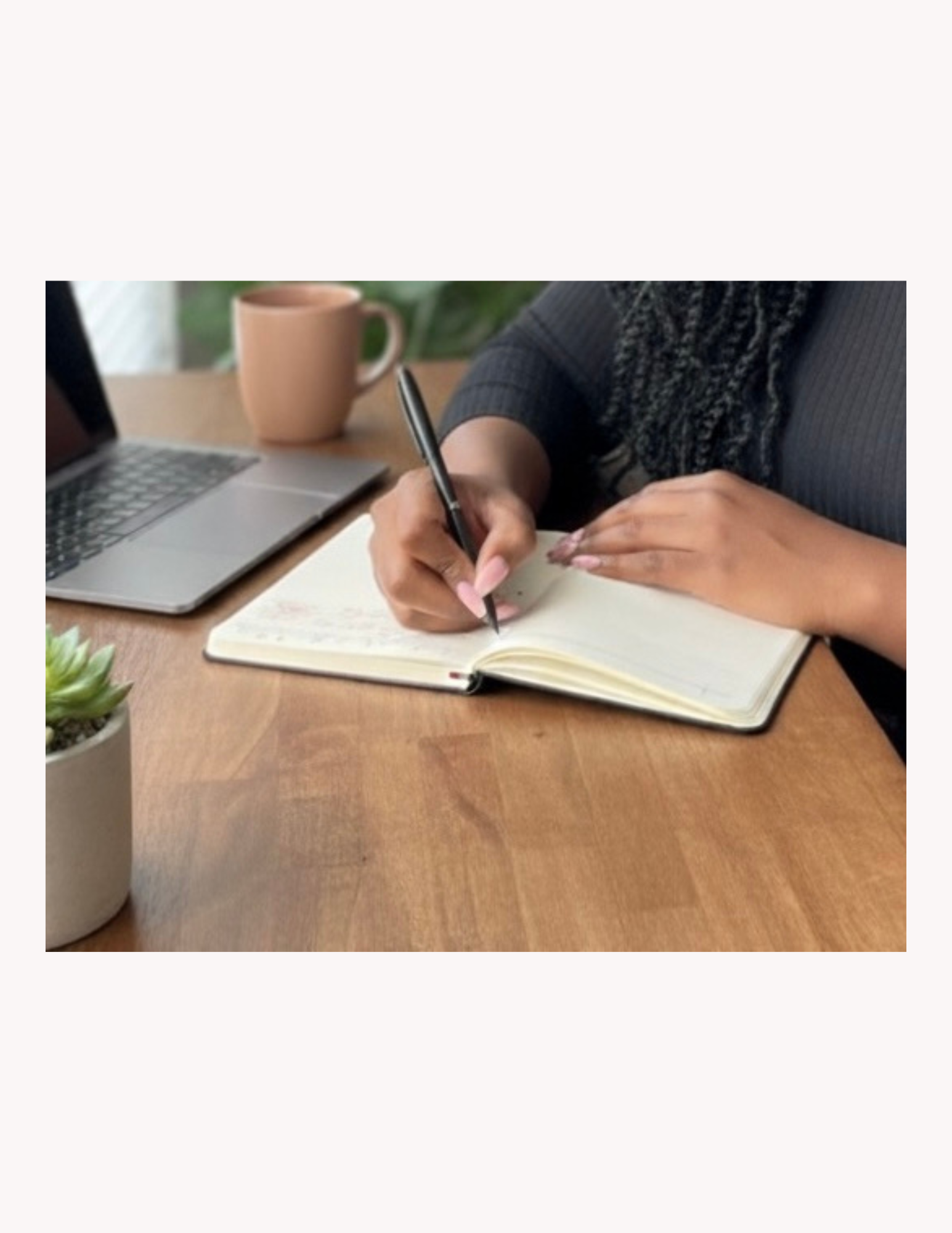 A person writing in a notebook at a wooden desk with a laptop, a mug, and a potted plant nearby.