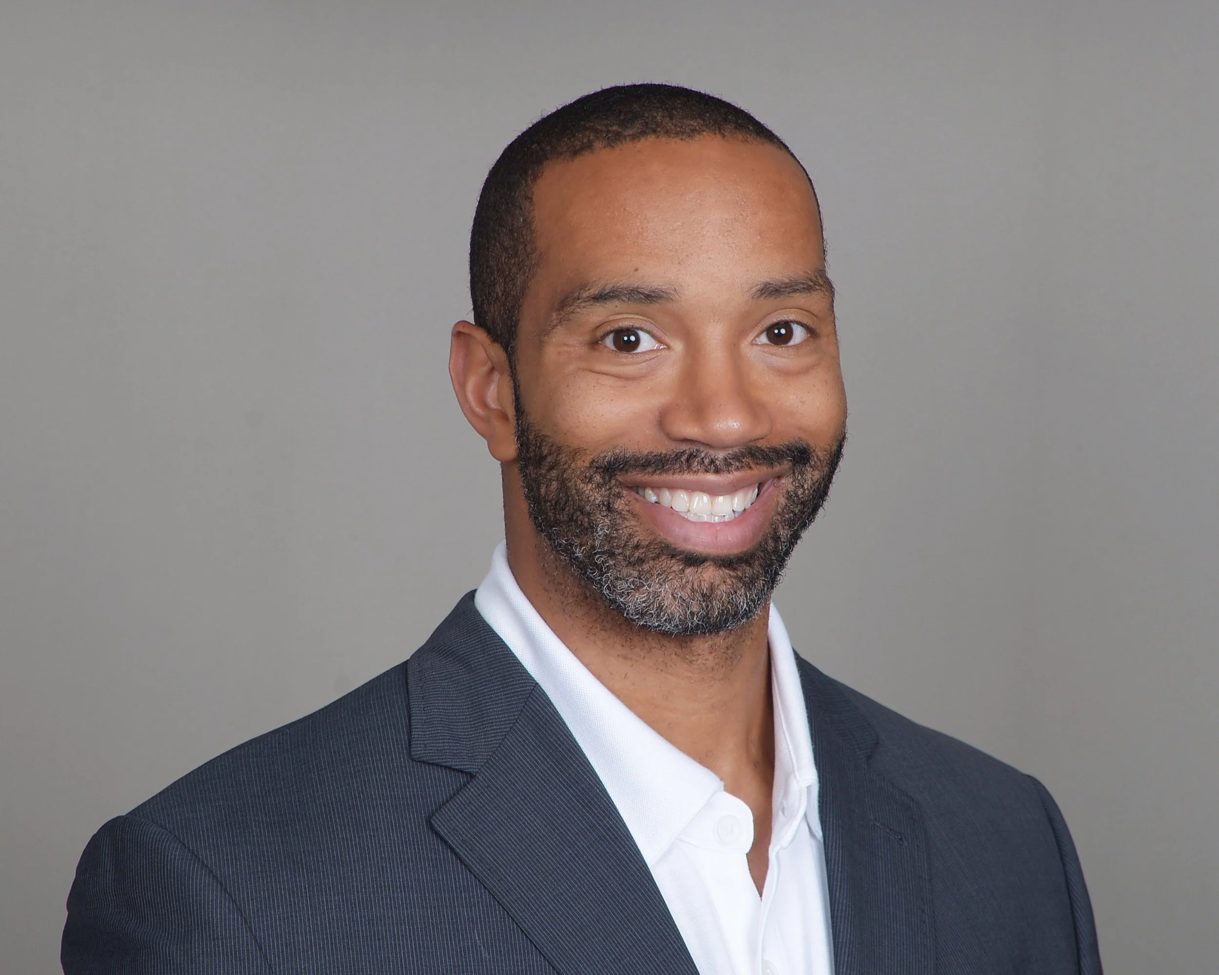 Headshot of a smiling African American man with short hair, beard, wearing a navy suit and white shirt, against a light gray background.