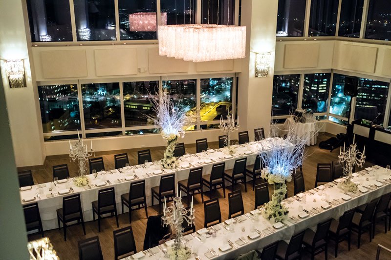 Elegant banquet hall with long tables set for a formal event, decorated with tall floral centerpieces and chandeliers, large windows showing city lights at night.