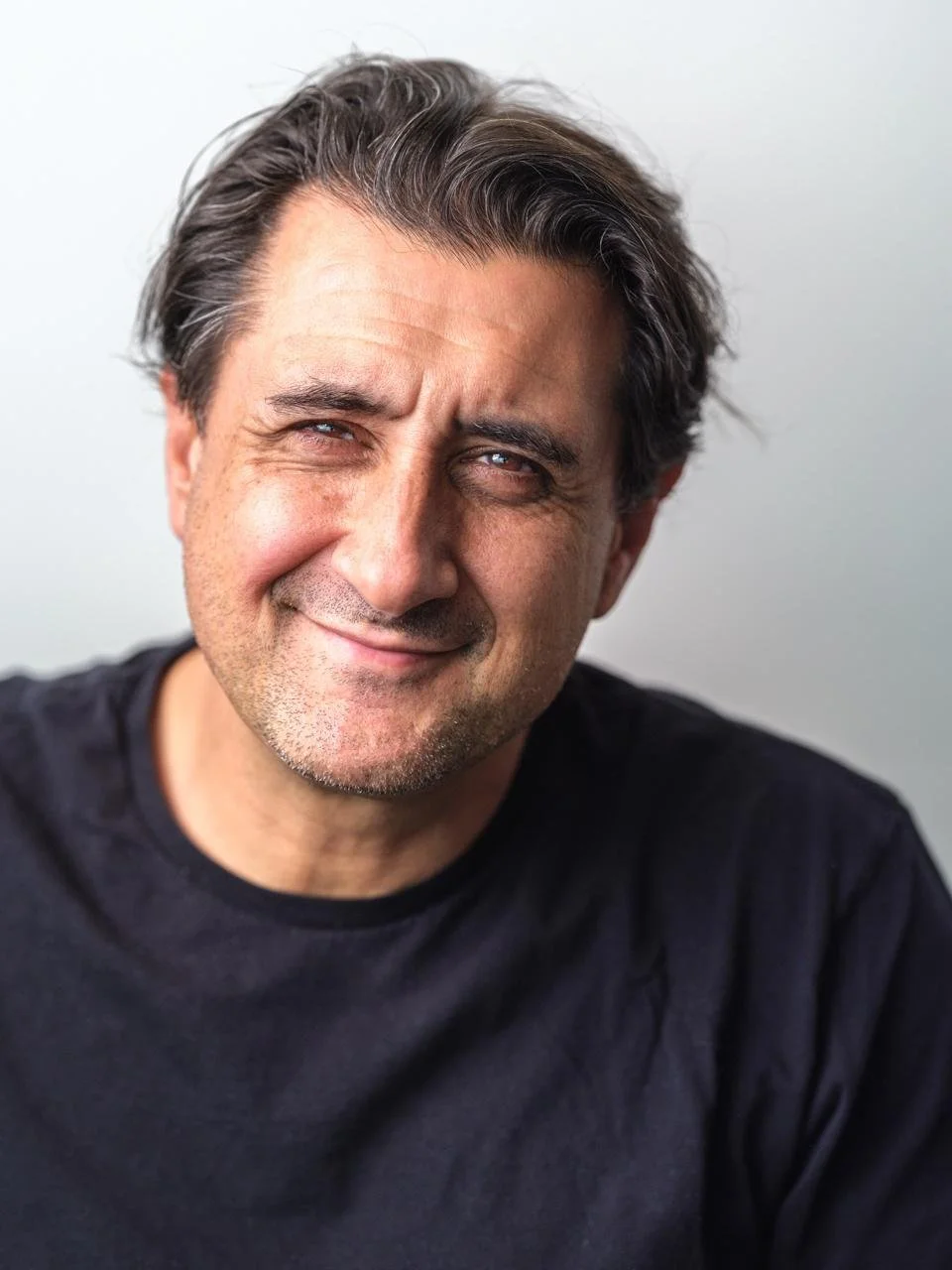 A middle-aged man with dark hair, slightly graying, smiling wearing a black shirt, with a light gray background.