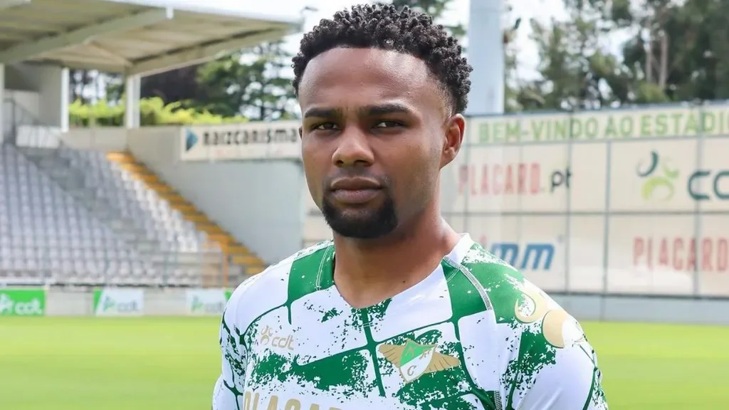 A young man with short curly hair and a beard, wearing a green and white sports jersey, standing on a football field with empty bleachers in the background.