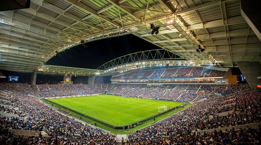 Nighttime view of a packed soccer stadium with a green field, spectators filling the stands, and a roof overhead with lighting.