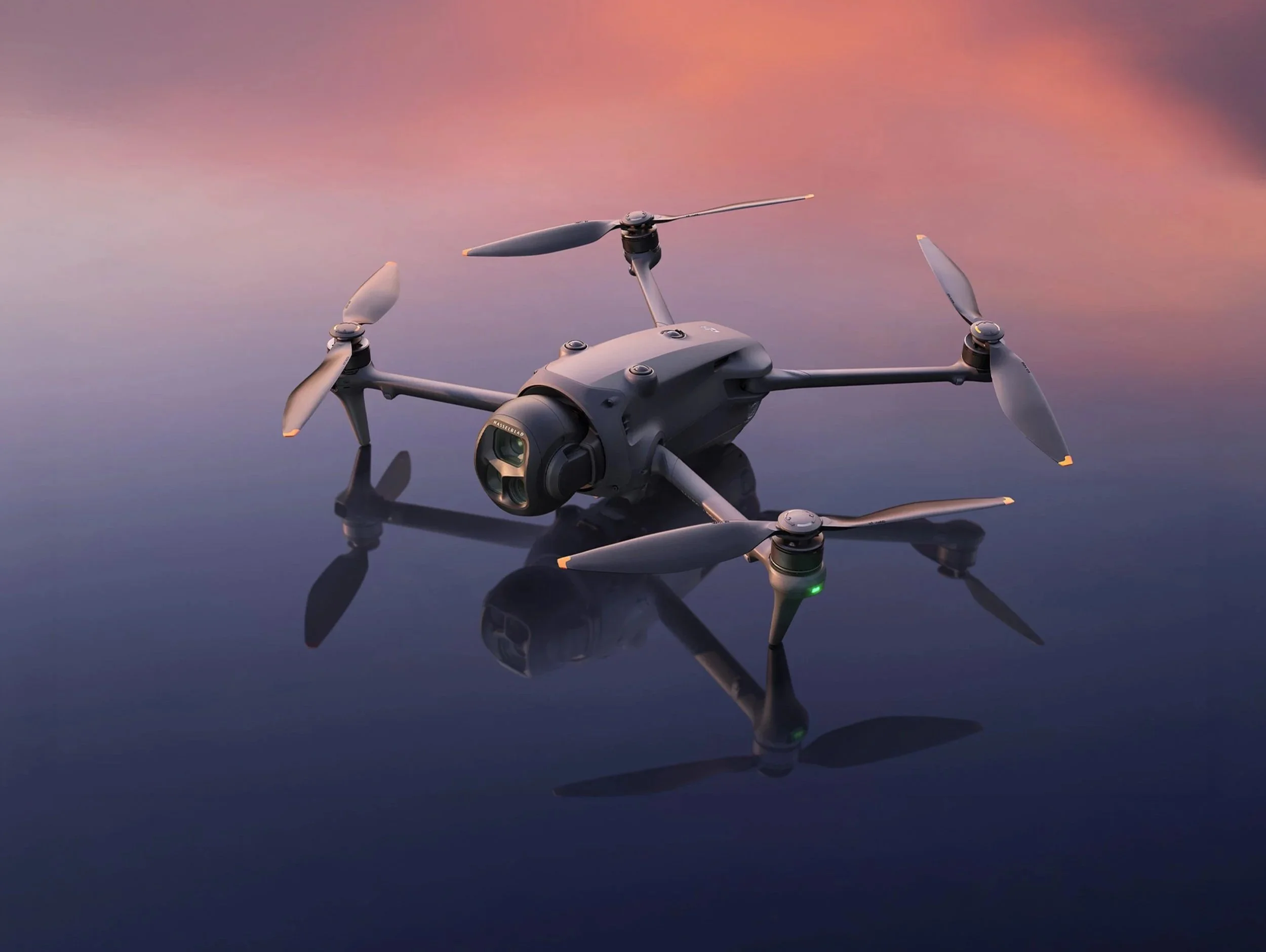 A black quadcopter drone with four propellers is positioned on a reflective surface during sunset or sunrise with a pinkish sky.