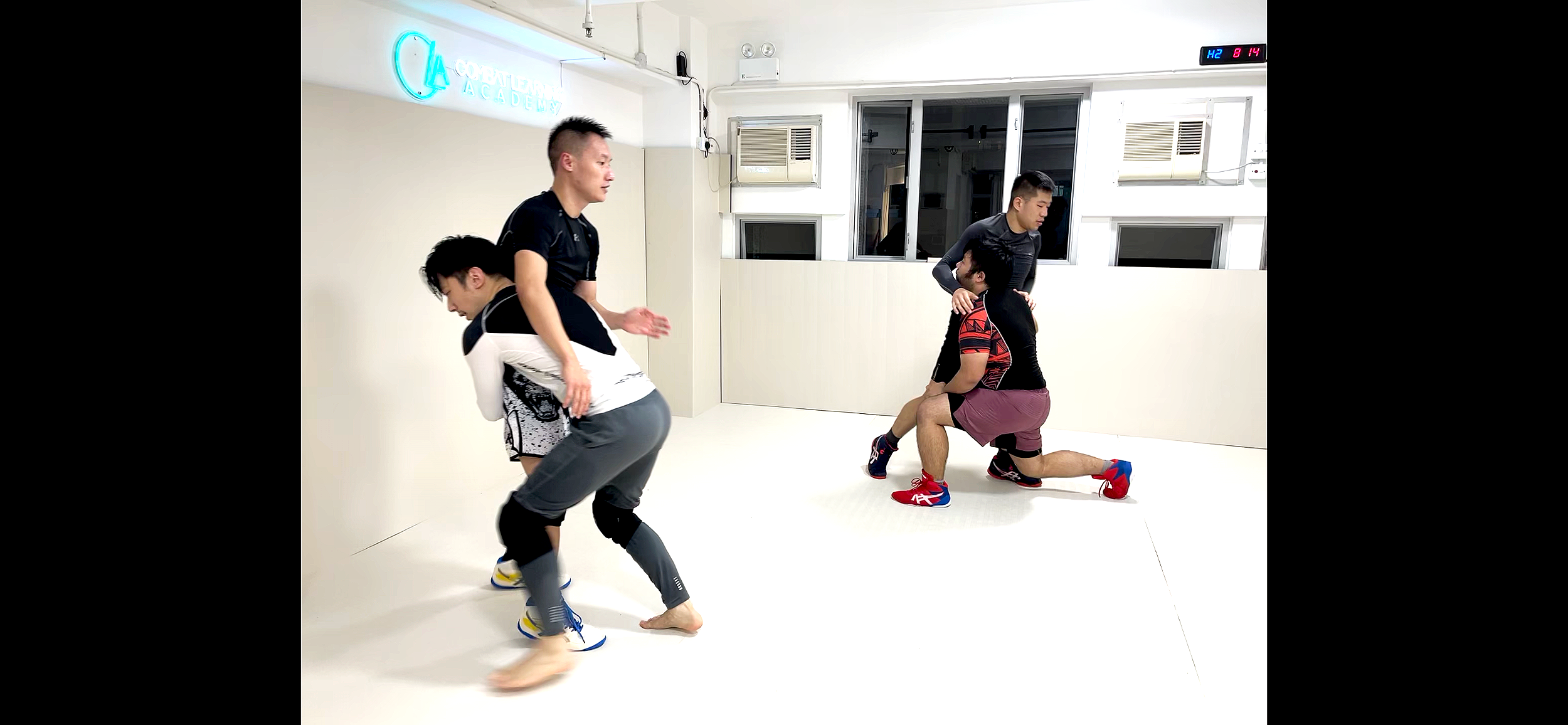 Two pairs of men engaged in a grappling or wrestling practice inside a gym. One man is kneeling and holding another from behind, while the other man is standing and appears to be preparing to defend or attack. The gym has white walls, windows, and a sign that reads 'Coconut Tree Academy' on the wall.