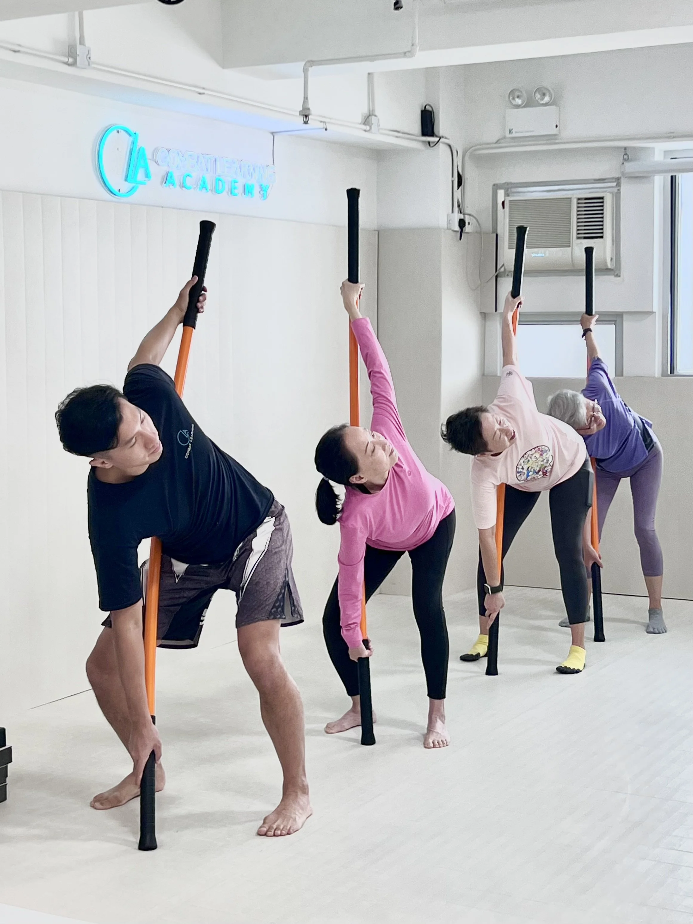 Group of people participating in a movement & mobility class, doing side stretches with mobility sticks, in a mirrored studio with a sign that says 'Combat Learning Academy' on the wall.