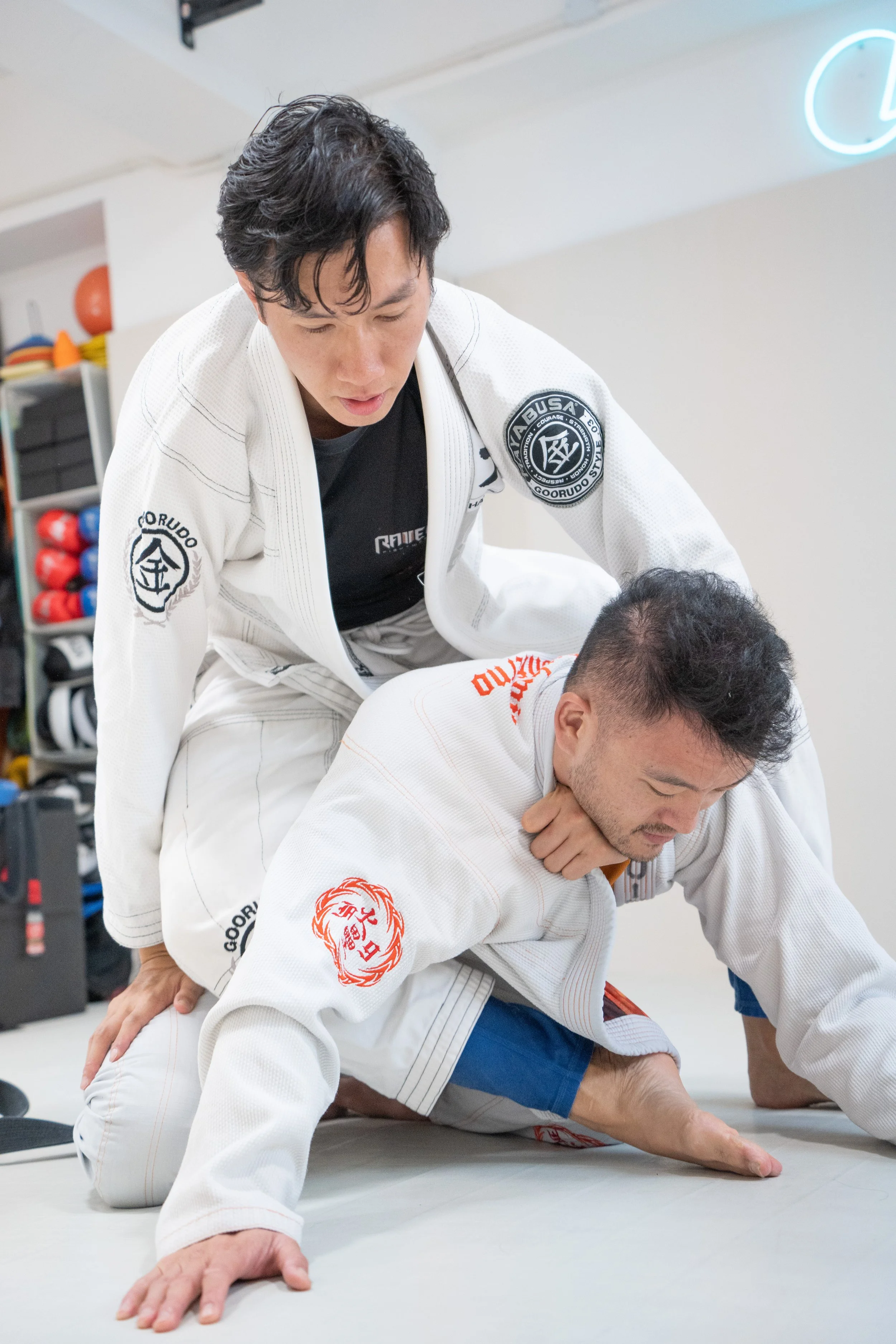Two martial artists practicing Brazilian Jiu-Jitsu on a mat indoors. One person is wearing a white gi with patches and is demonstrating a technique on another person also in a gi. The background has gym equipment and a neon sign.
