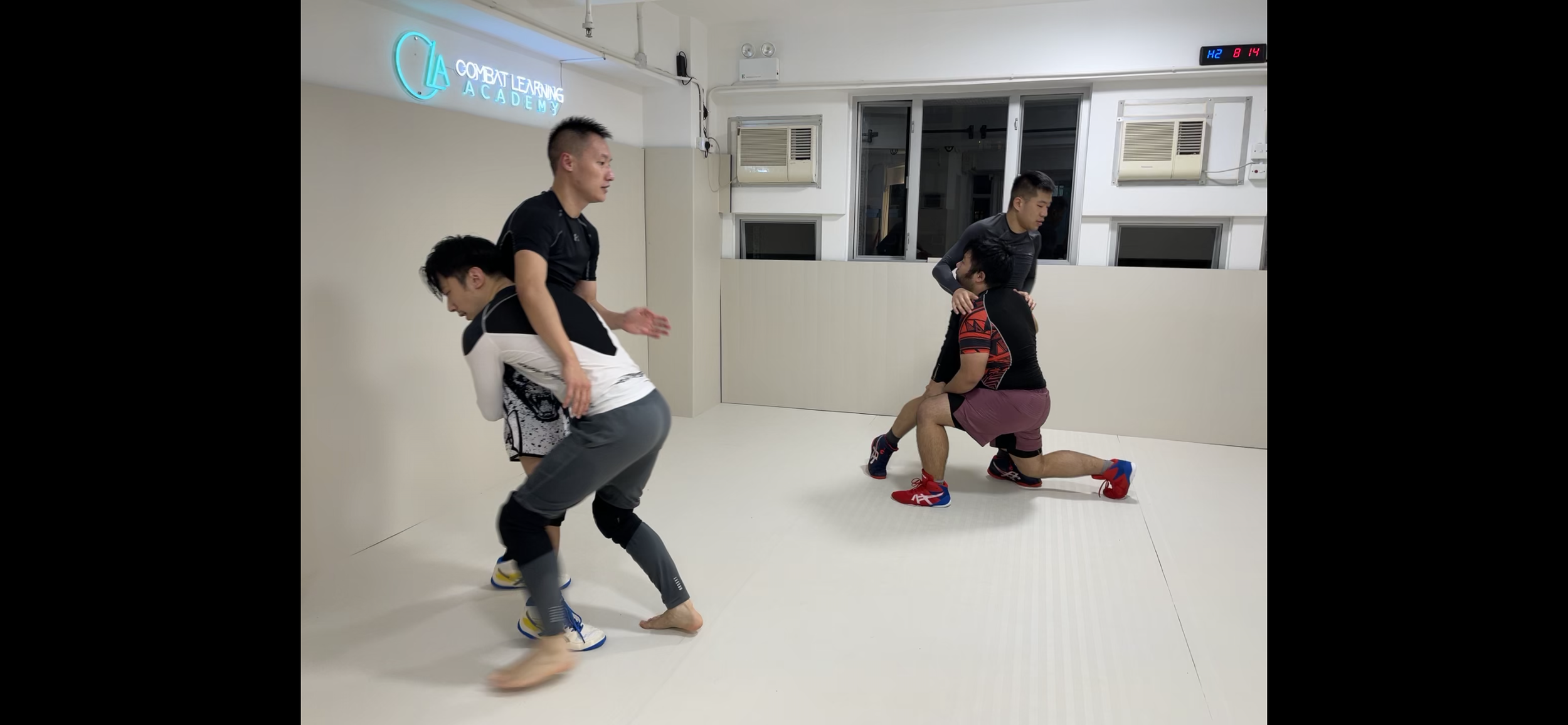 Four men practicing wrestling in a gym with a white mat. Two men are in the foreground, one in a black shirt and maroon shorts kneeling, the other in a white and black shirt. A man in a black shirt is standing behind them, and another man is standing further back in a black shirt. The gym has a sign that says 'Combat Learning Academy' on the wall, and a digital clock showing the time as 8:14.