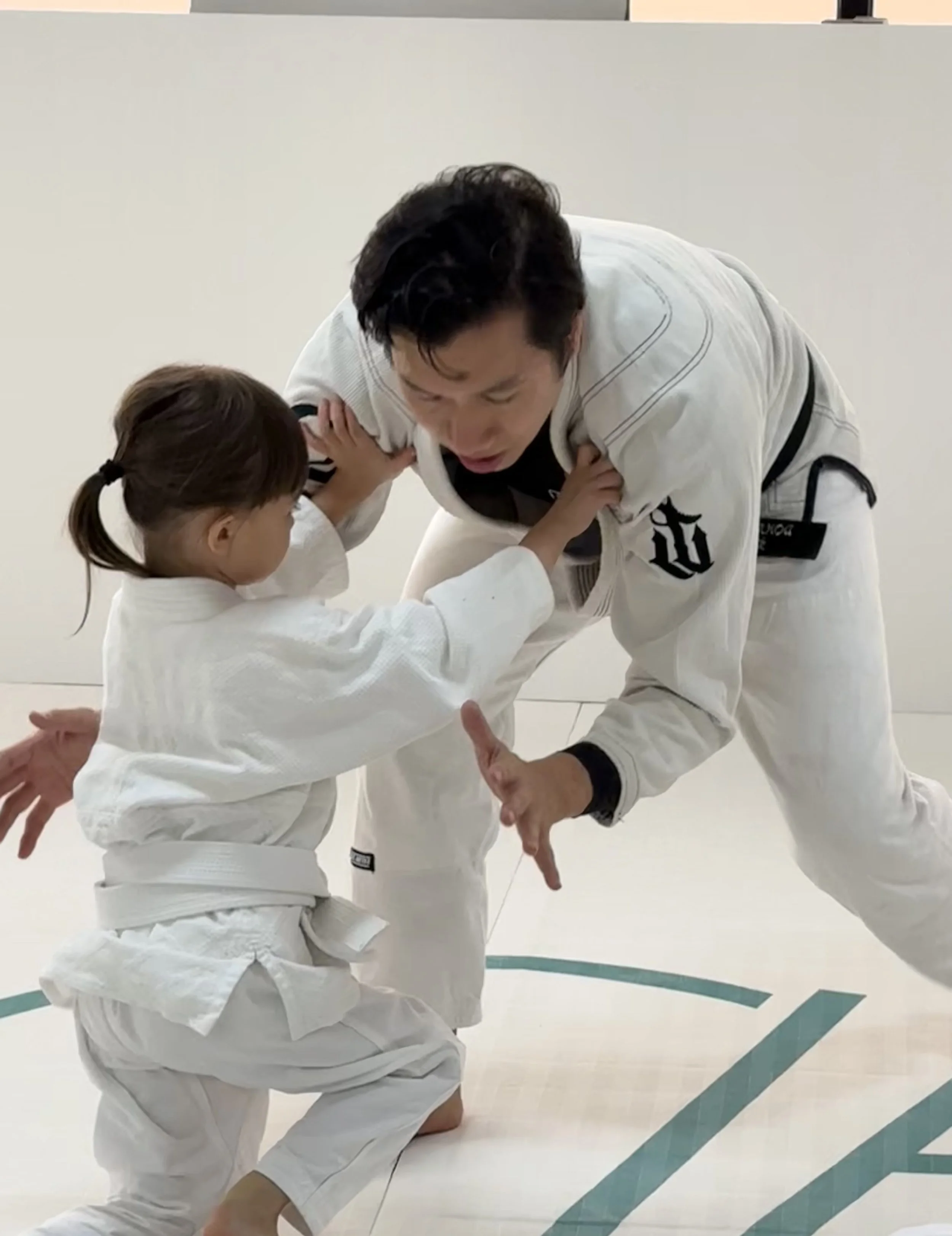 An adult and a child practicing judo in a dojo, wearing white gi uniforms. The adult appears to be teaching or demonstrating a hold or move, with the child actively engaging in the practice.