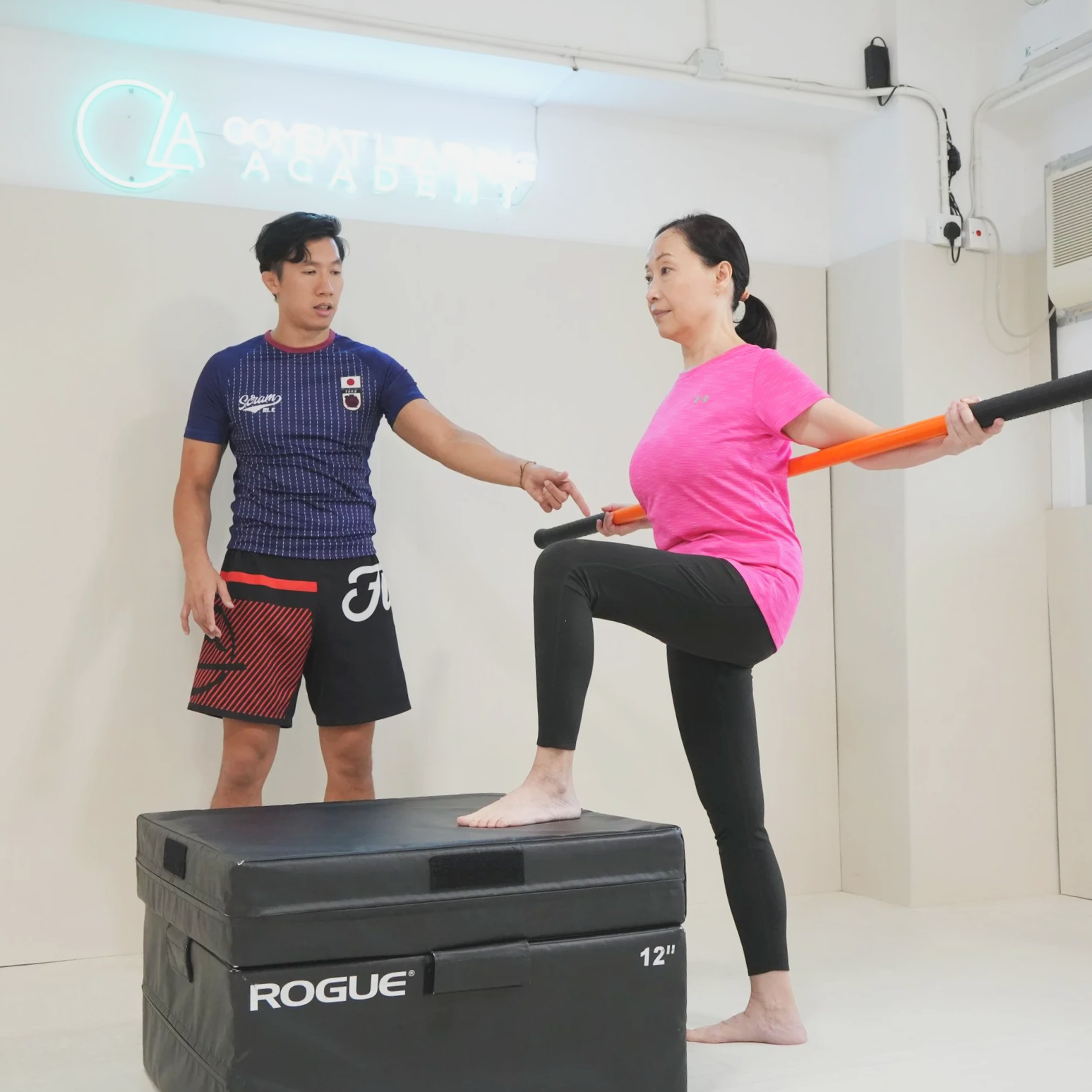 A woman is practicing balance exercises on a padded platform with the help of a trainer in a gym. She is holding a mobility stick behind her shoulders and balancing on one foot, while the trainer observes and points at her.