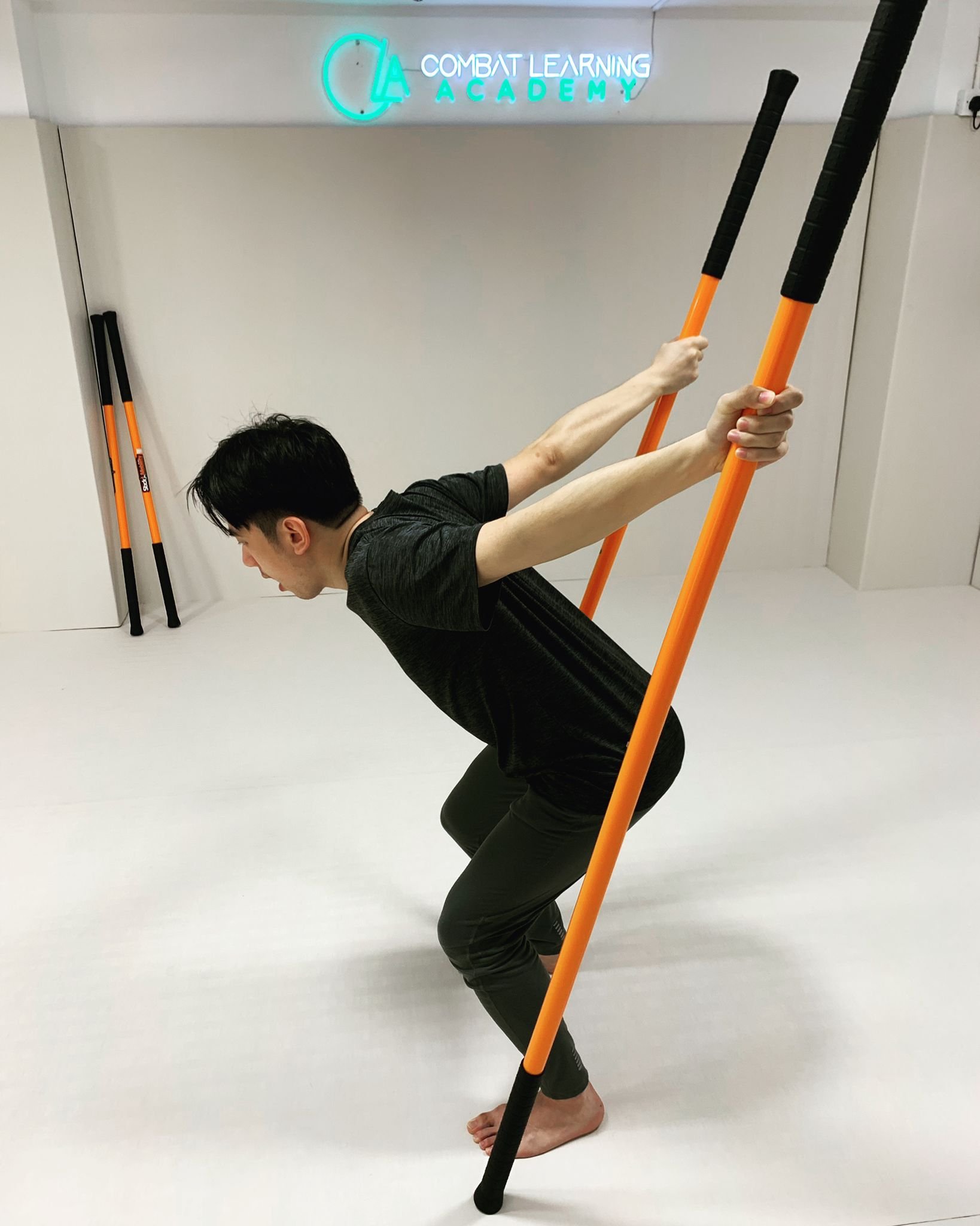 A person practicing a yoga pose with orange and black sticks in a room with a sign that reads "Combat Learning Academy" on the wall.