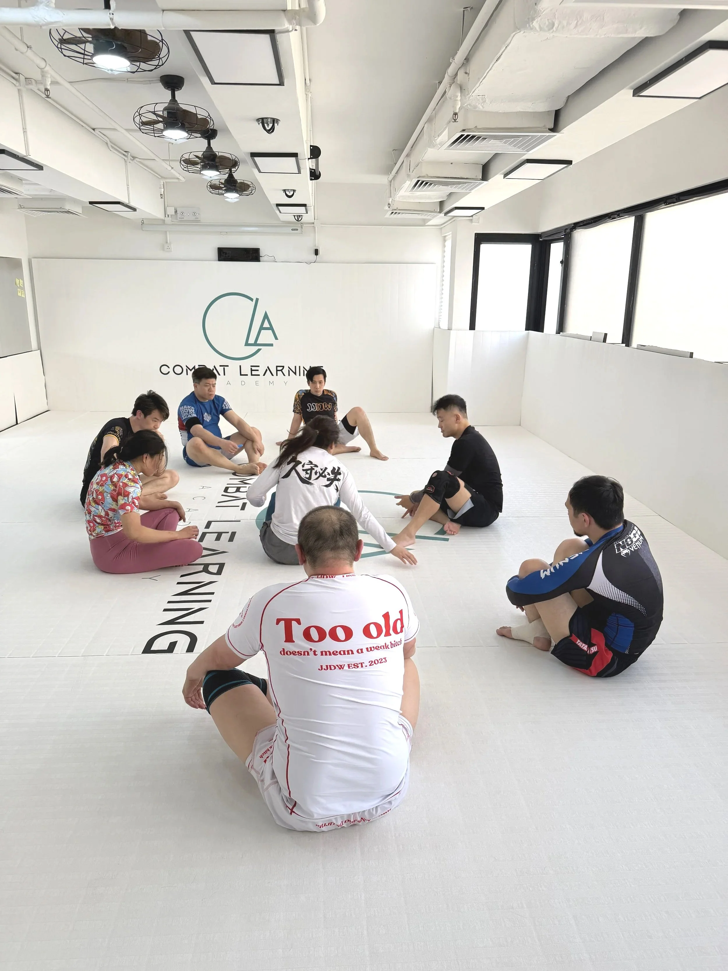 Group of people sitting in a circle in a martial arts training room with white walls and mats, some wearing casual clothes and others in sportswear, engaging in a discussion or instruction session.
