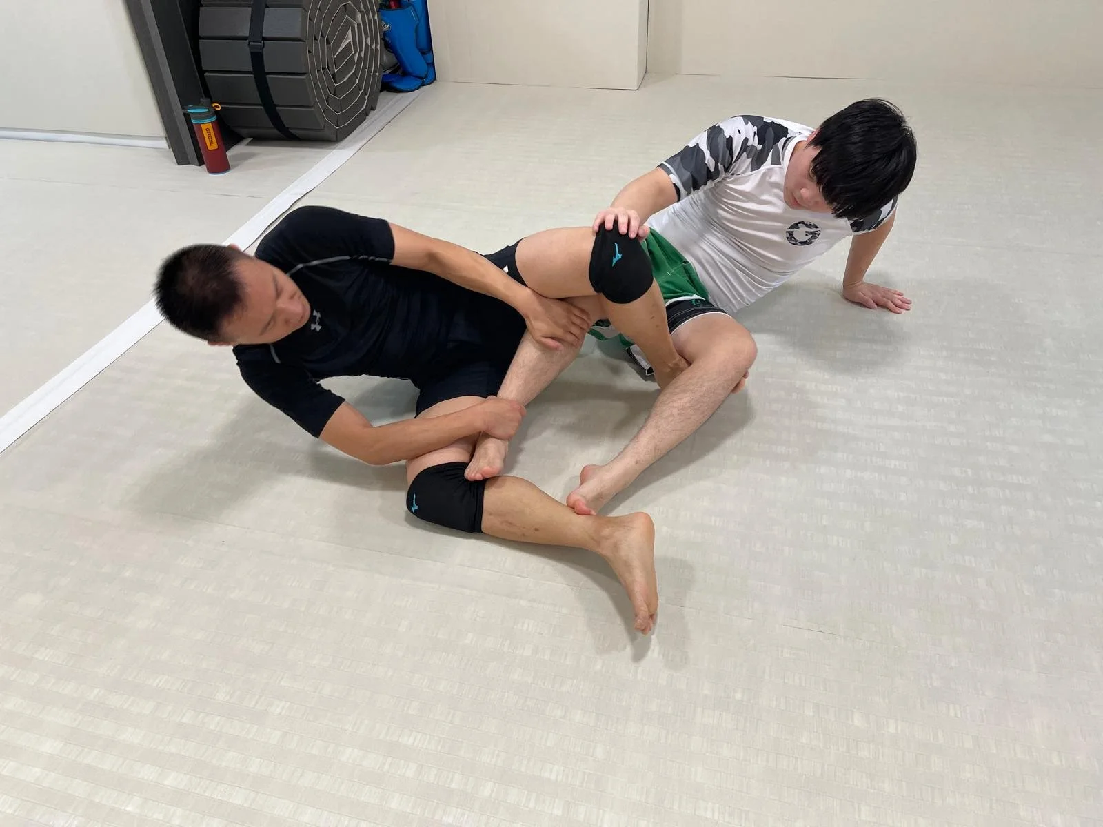 Two men practicing Brazilian Jiu-Jitsu on a light-colored mat. One man is on his back and the other is on top, both wearing athletic rash guards and shorts. The man on top has his hands on the other man's leg while the man on the bottom is holding his opponent's arm and appears to be in a defensive position.
