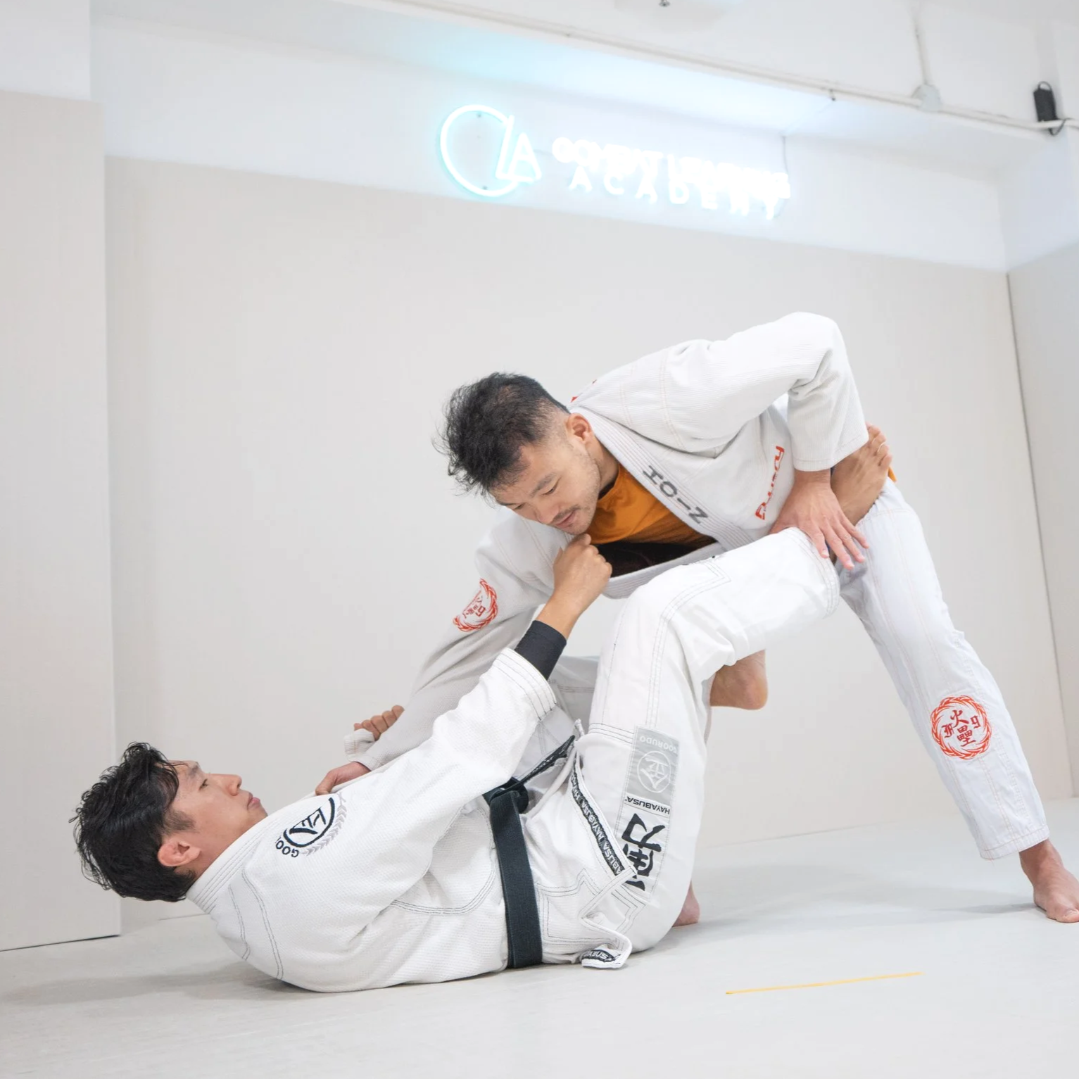 Two men dressed in white Brazilian Jiu-Jitsu gis practicing a grappling move in a martial arts gym, with one man on the ground and the other standing over him, both focused on their technique.