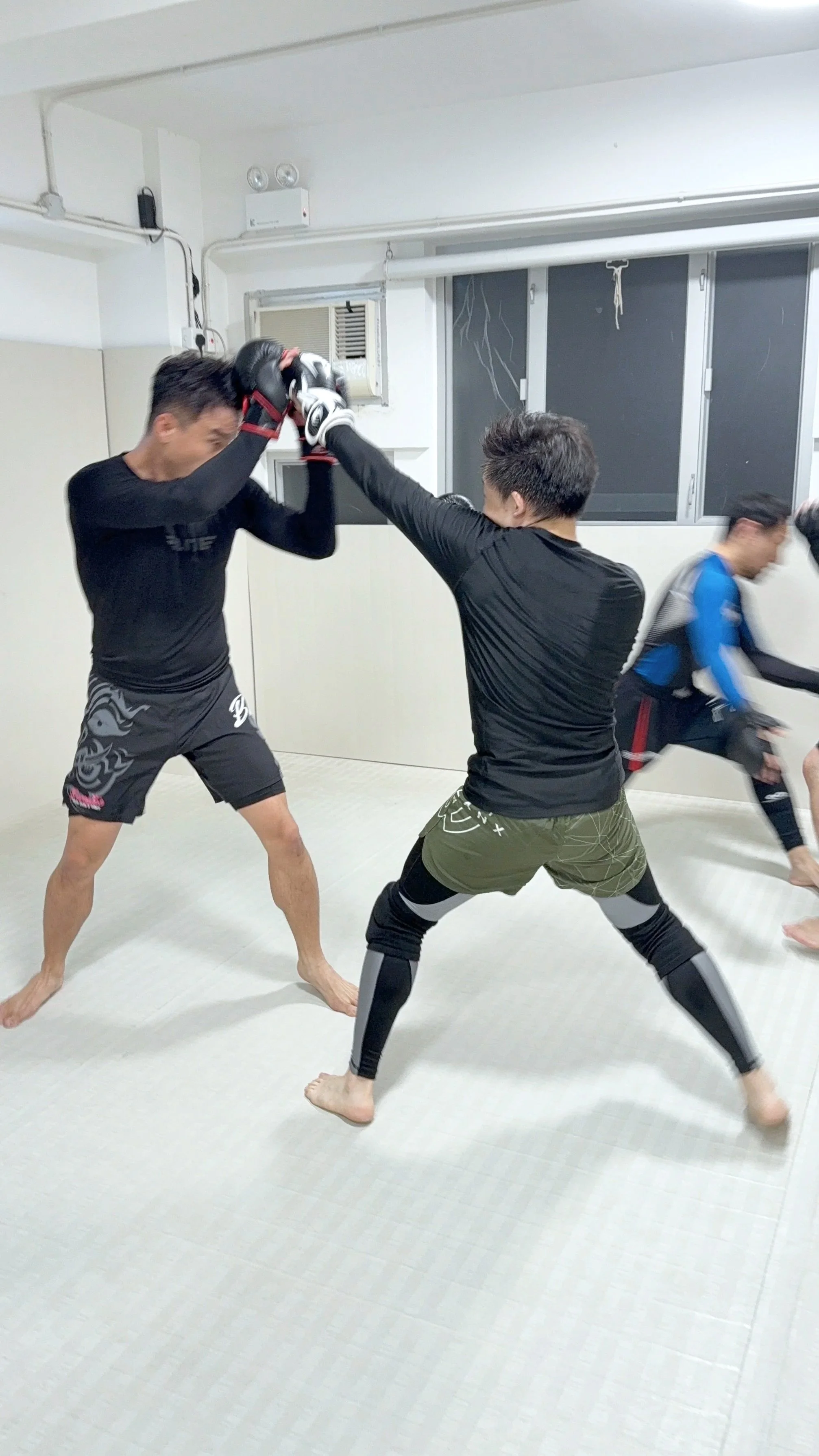 Two martial artists practicing sparring with gloves in a training room, while a third person is in the background.