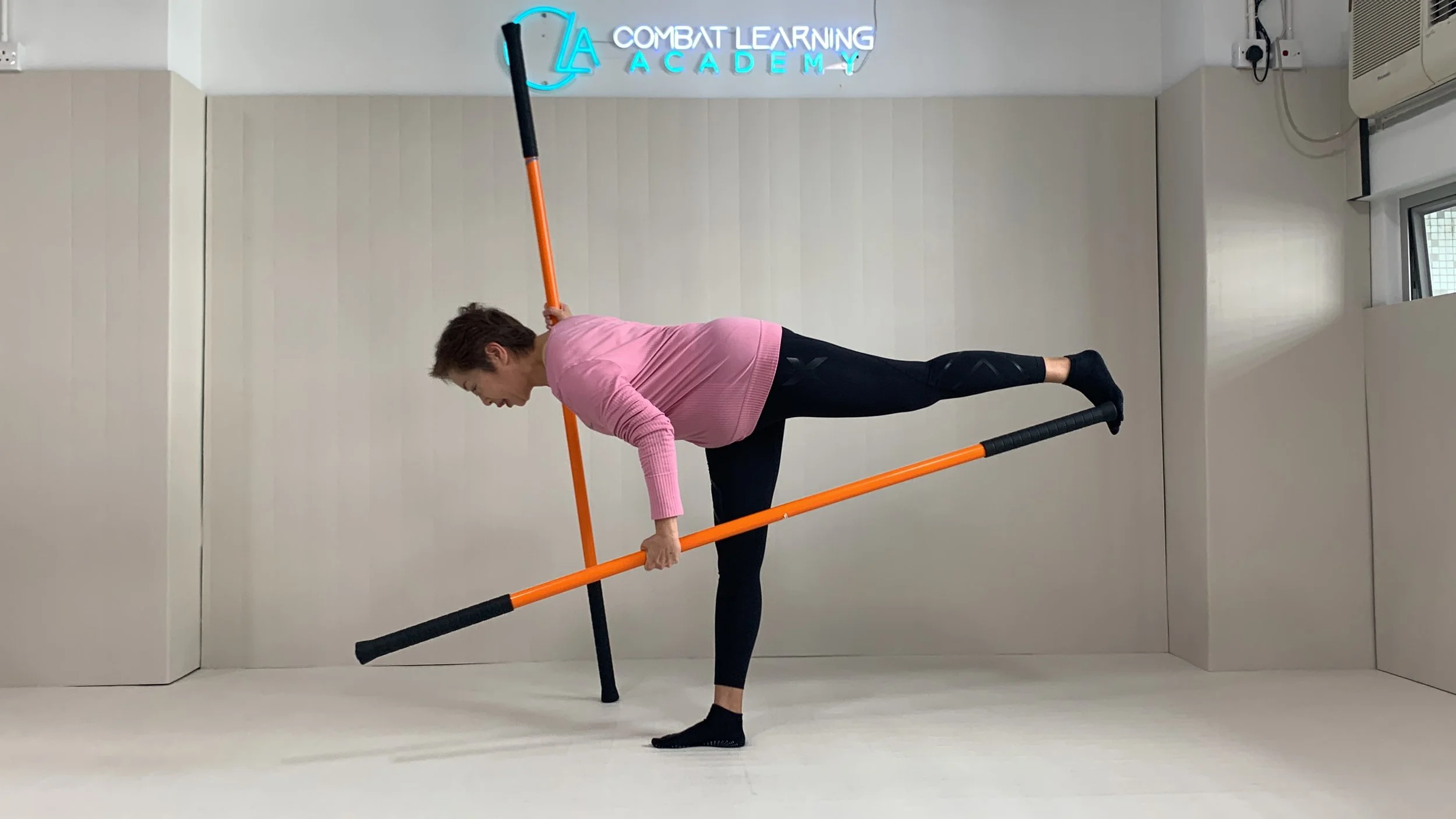 A woman in a pink long sleeve top and black leggings practicing a yoga pose with orange and black sticks in a room with white walls. There is a neon sign on the wall that reads "LA Combat Learning Academy."