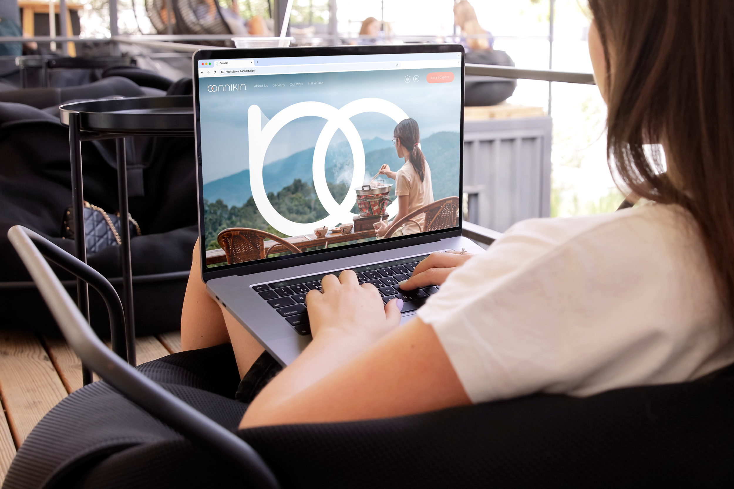 A person sitting in a cafe using a laptop, which displays a website with a scenic background of mountains and a woman drinking tea or coffee. The cafe has wooden flooring and other people in the background.