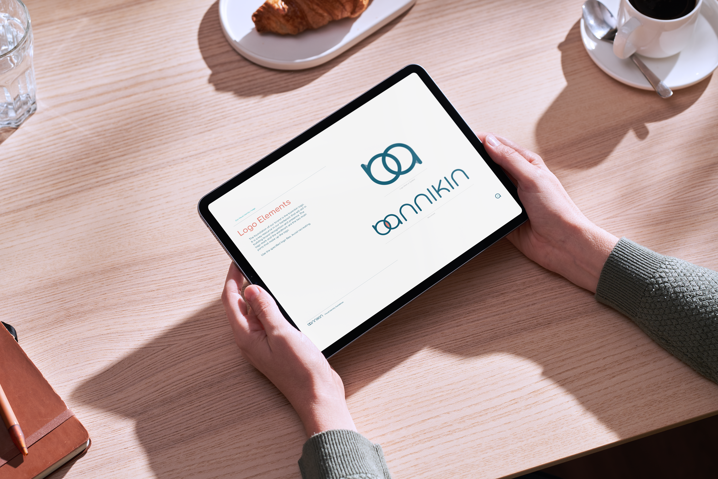 Person holding a tablet displaying a logo design presentation with a wooden table, cup of coffee, croissant, glass of water, and notebook nearby.