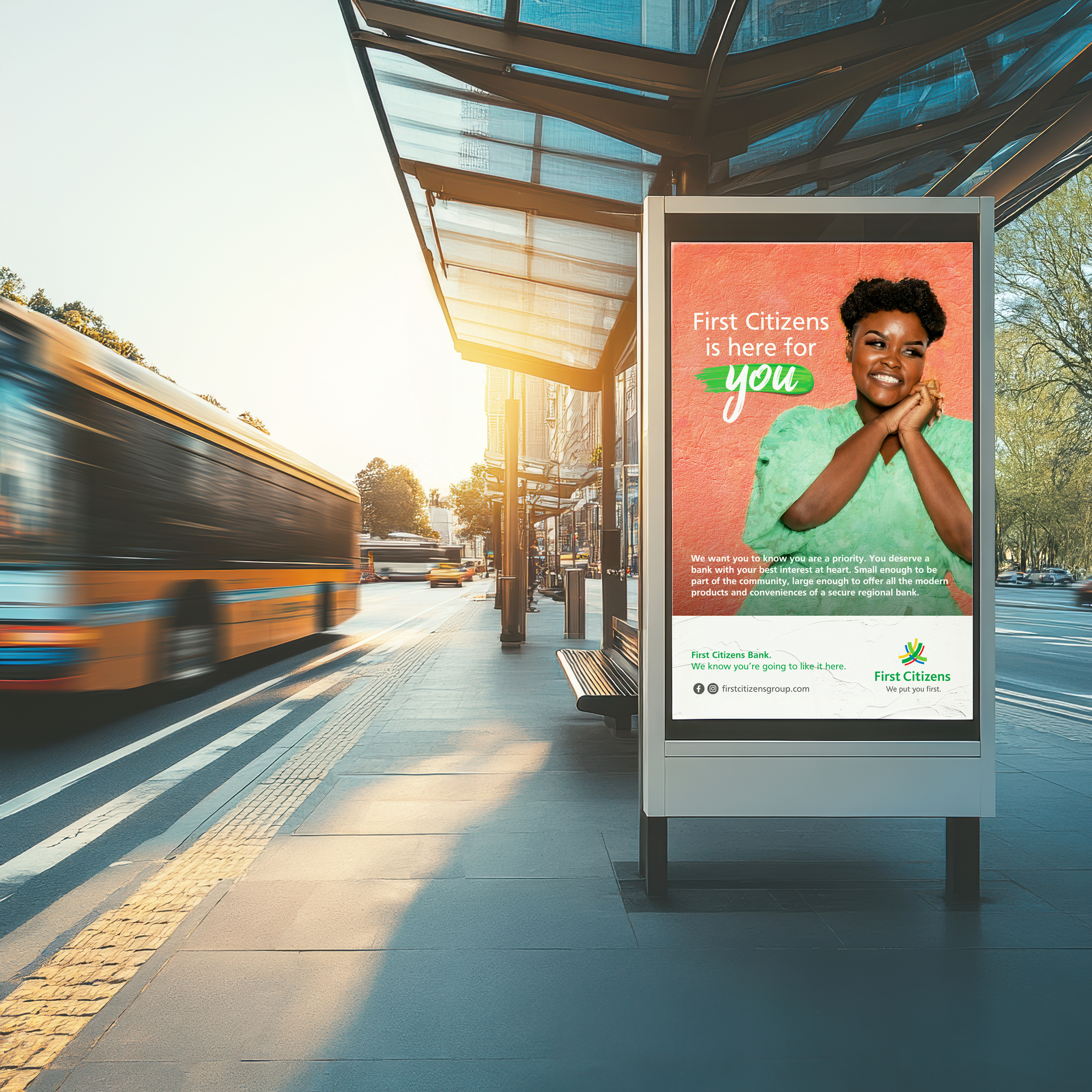 A digital billboard at a bus stop on a city street in the late afternoon or early evening. The billboard displays an ad for First Citizens Bank with a smiling woman and the text "First Citizens is here for you." There is a yellow bus approaching in the background and the bus stop shelter features a modern glass roof and a bench.