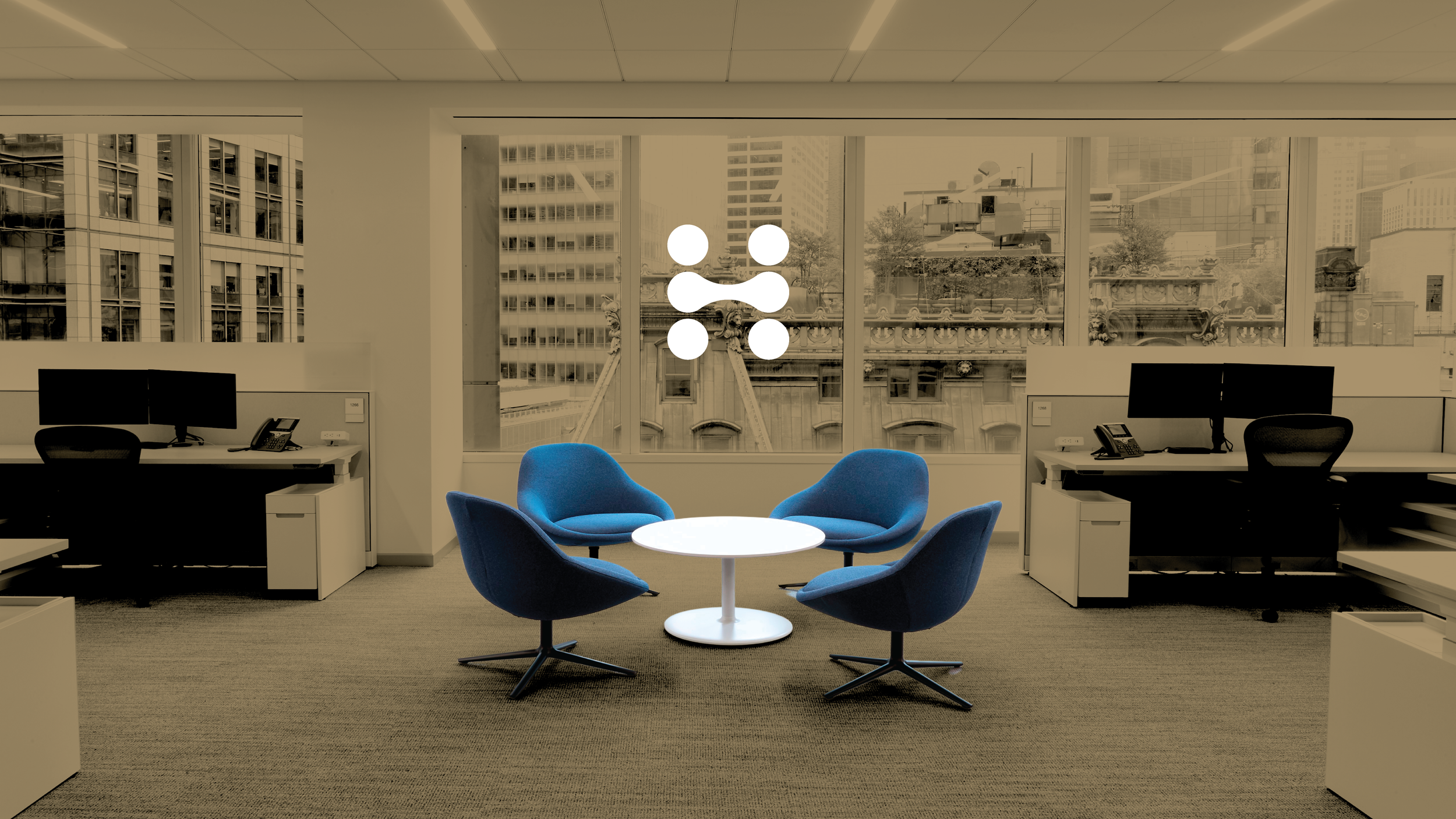 Modern office with two desks and two computer monitors on each, a round white table with four blue chairs in the center, large windows showing city buildings, and a logo in the window with four circles and a connecting line.