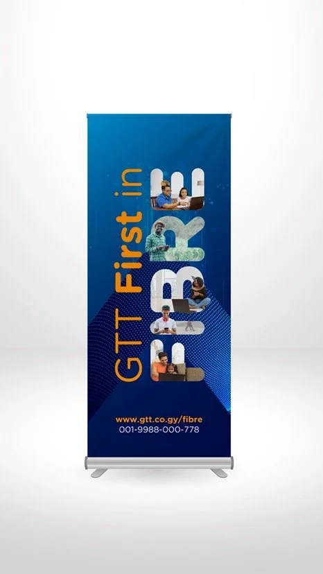A vertical blue roll-up banner promoting GTT's fiber internet service. It features the text 'GTT first in FIBRE' and includes images of diverse people using technology, with the website www.gtt.co.gv/fibre and a contact number at the bottom.