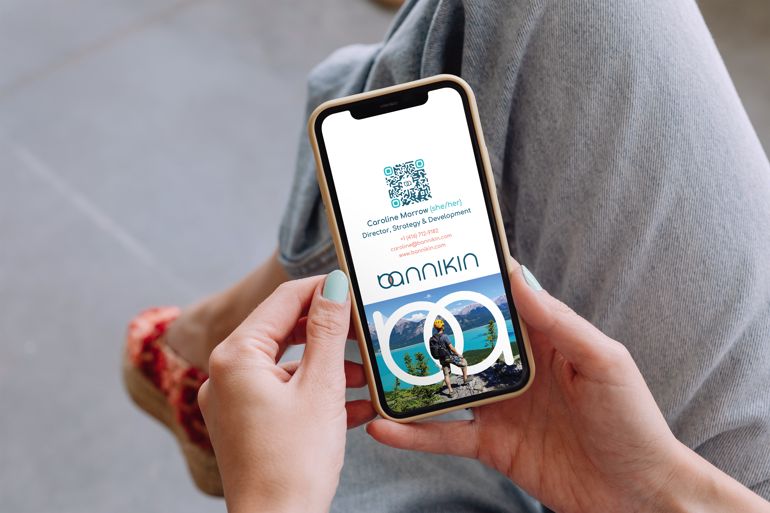Close-up of a person holding a smartphone displaying a digital business card for Caroline Morrow, Director of Strategy & Development, with a scenic mountain lake background and a QR code.