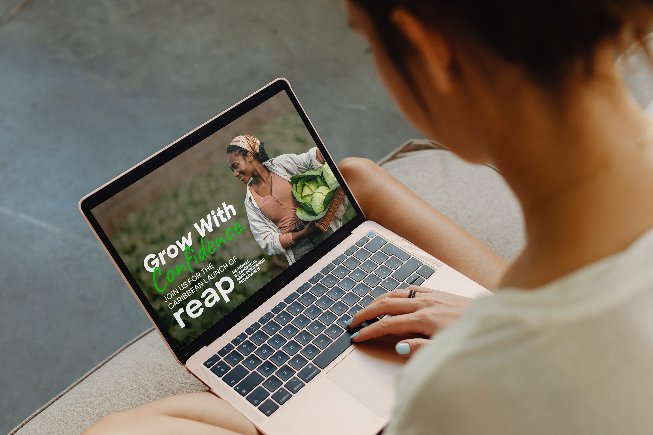 A woman sitting on a couch using a laptop that displays a promotional poster for a Caribbean agricultural program, featuring a smiling woman holding a basket of fresh vegetables with the text 'Grow With Confidence' and details about joining the launch.