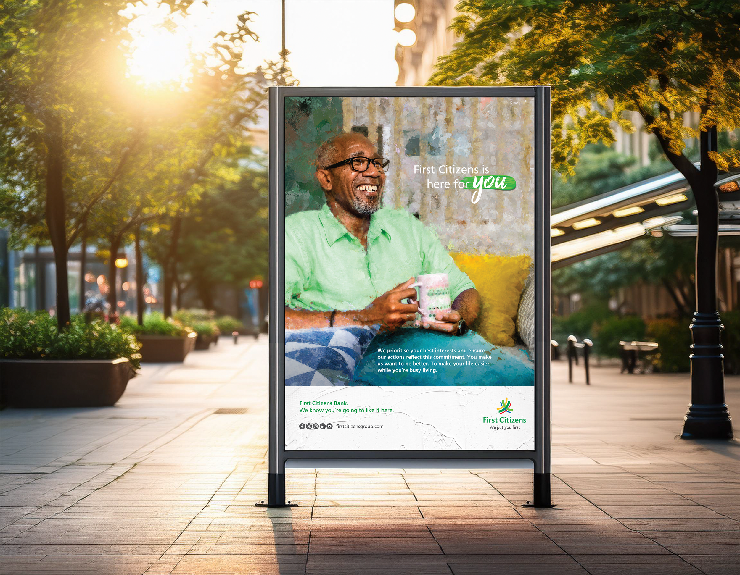 An outdoor sidewalk advertisement for First Citizens Bank featuring a smiling man with glasses in a green shirt holding a mug, with text promoting the bank's services and slogan, shot during sunset on a tree-lined street.