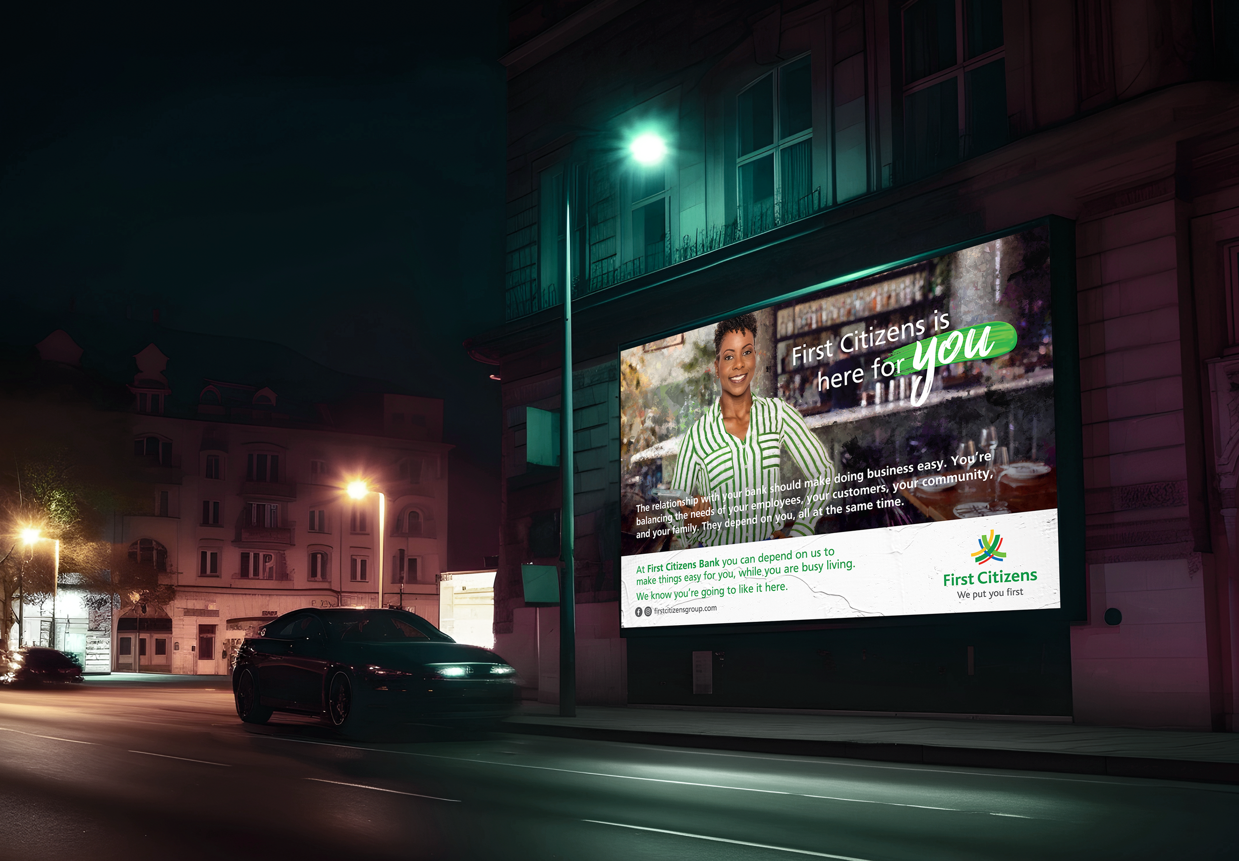 Night scene of a city street with a large illuminated billboard on the side of a building. The billboard features a smiling woman in a green and white striped shirt, advertising First Citizens Bank. The area has streetlights, a nearby car, and residential buildings in the background.
