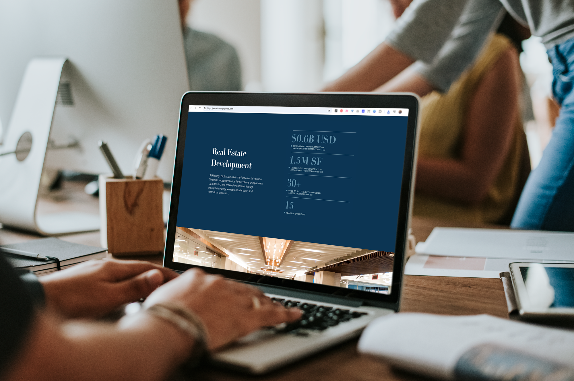 Laptop on a desk showing a real estate development presentation with statistics, surrounded by notebooks, papers, and office supplies in a workspace.