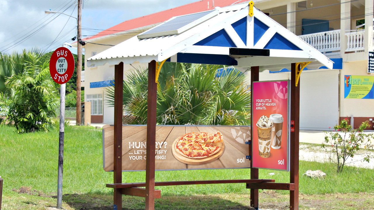 An outdoor advertisement shelter with a wooden frame, white roof, and blue gable accents, displaying a pizza and beverage ads, next to a bus stop sign that reads "BUS OUT OF CITY STOP," on a grassy area with trees and a building in the background.
