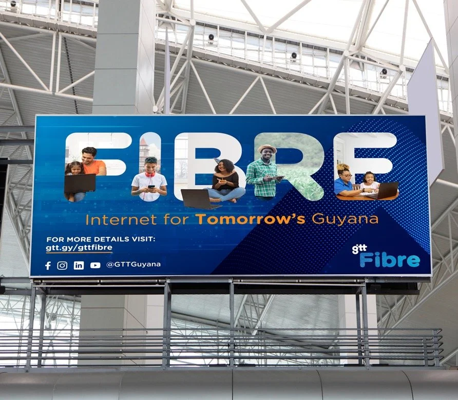 Large advertisement for fiber internet in Guyana with five diverse people using laptops and mobile devices, slogan 'Internet for Tomorrow's Guyana,' and social media icons.