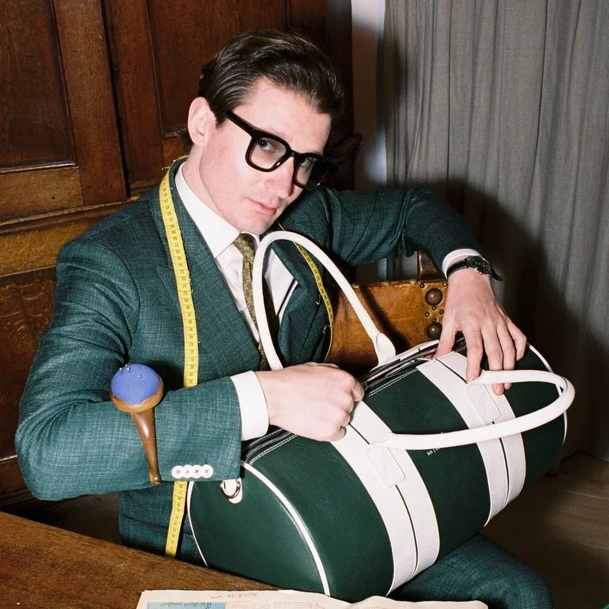 A man wearing glasses, a suit, and a measuring tape around his neck, sitting at a wooden table with a large black-and-white striped bag, holding an ice cream cone in one hand.