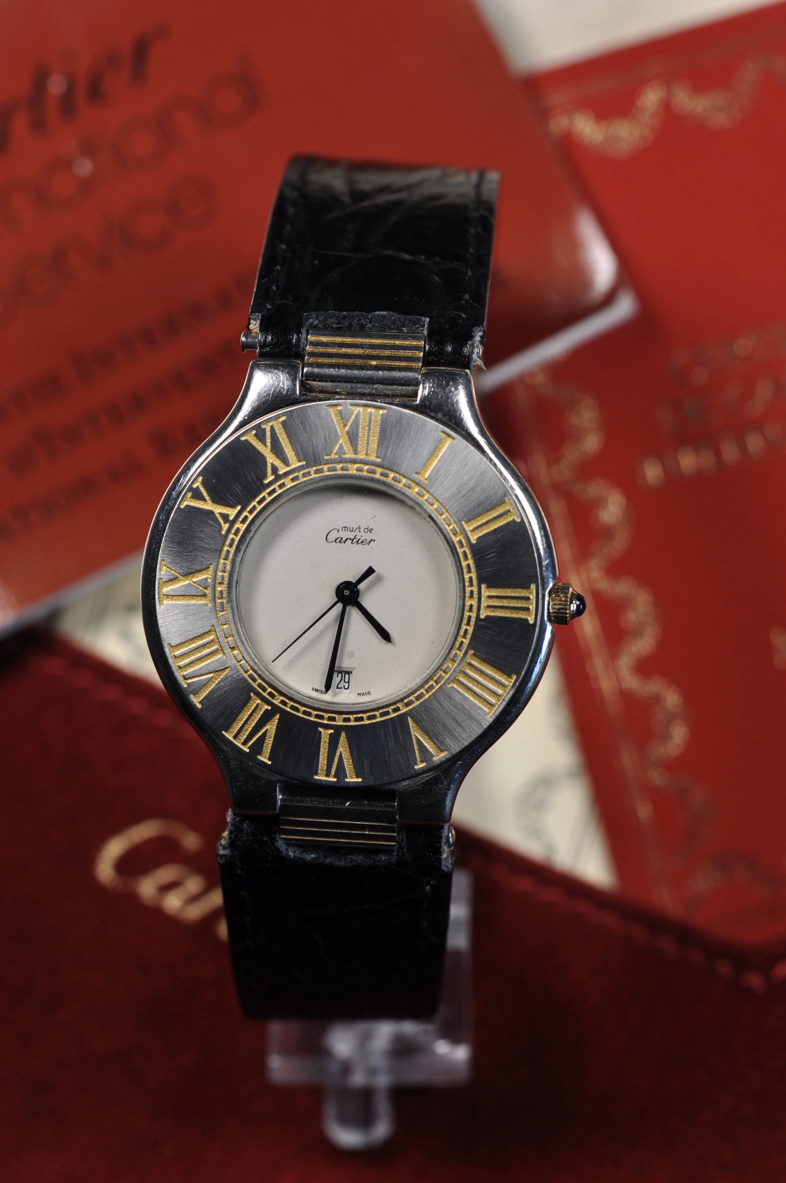 Close-up of a luxury wristwatch with a black leather strap, silver and gold dial, Roman numerals, and a date display, placed on a red jewelry box.