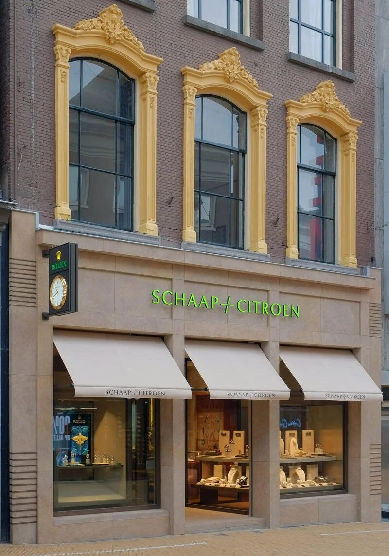 A storefront with a sign reading 'SCHAAP + CITROEN'. The building has three large windows with ornate yellow decorative framing. There is also a Rolex clock sign on the left side of the store.