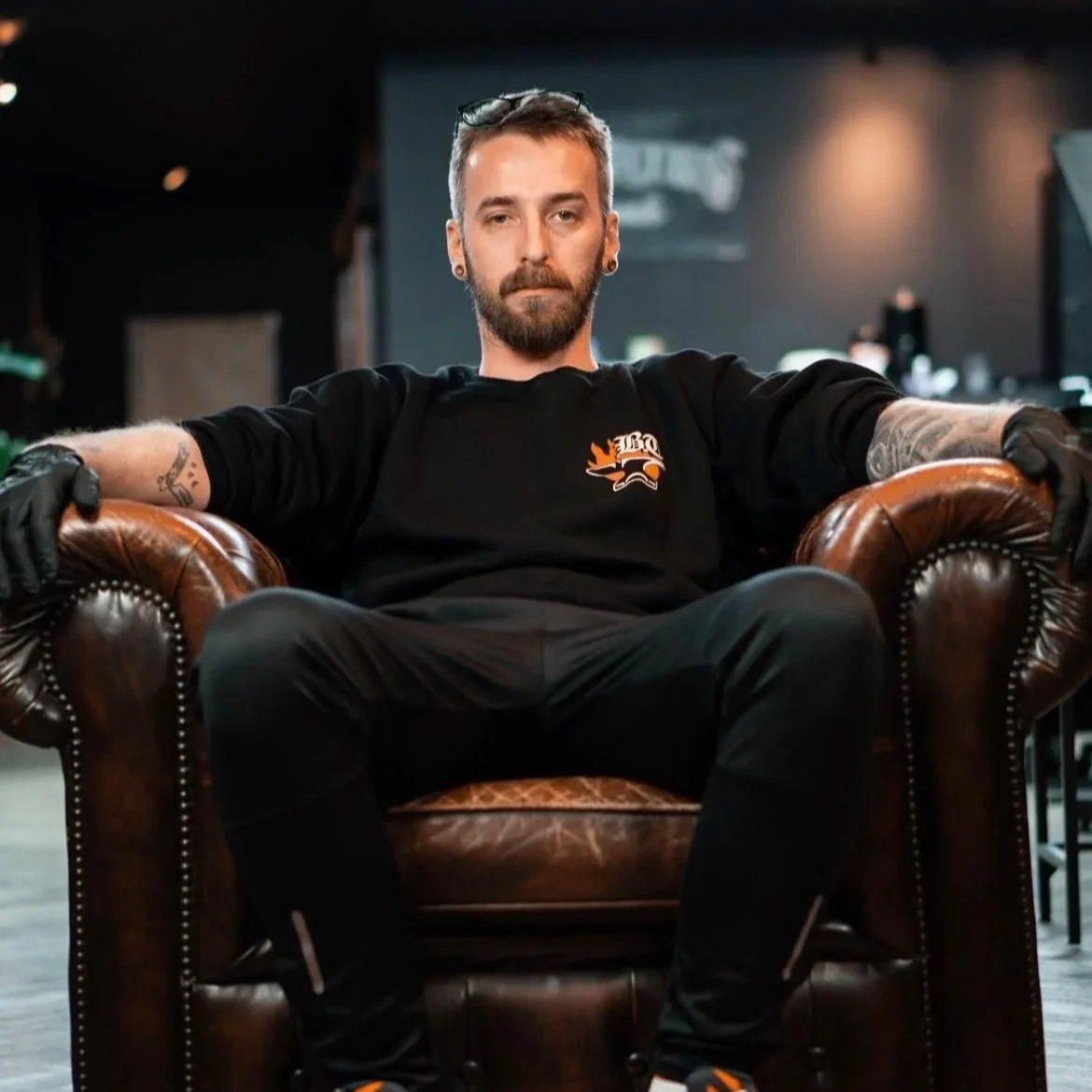A man with tattoos and facial piercings sitting confidently in a leather armchair in a bar or lounge setting, wearing a black shirt with logo and black gloves.