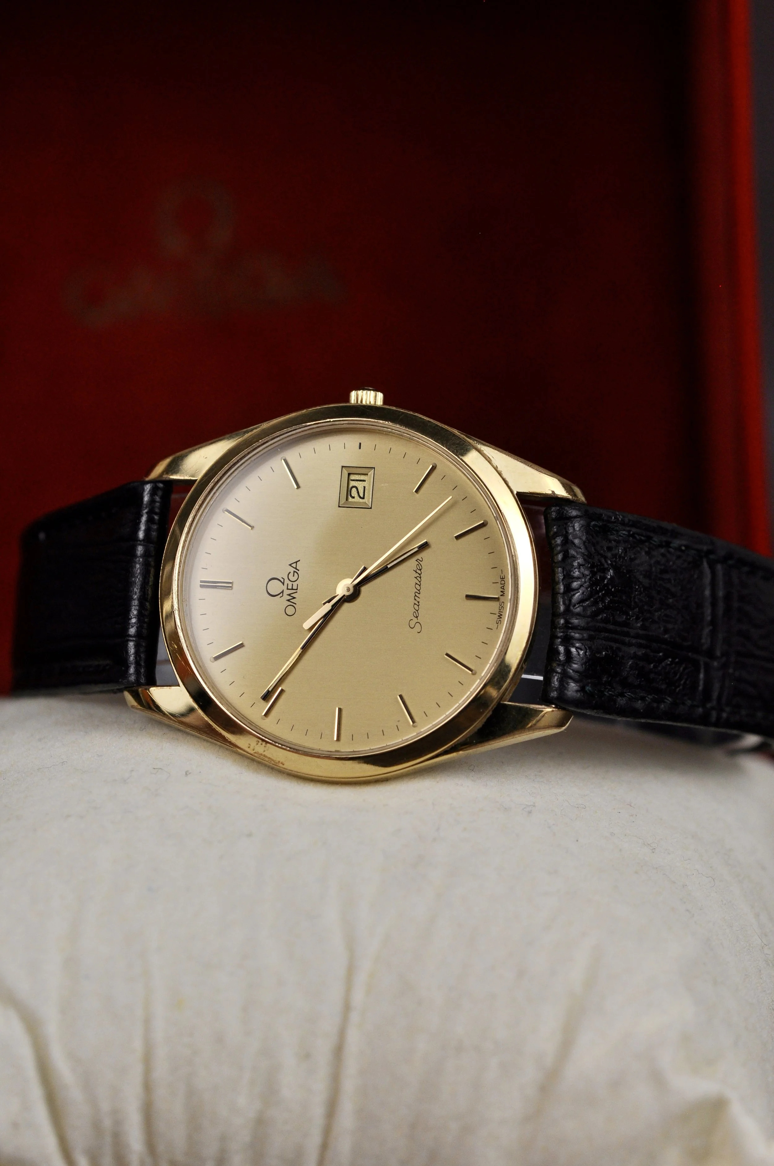 Gold Omega Seamaster wristwatch with a beige face, black leather strap, and date display, resting on a white surface with a blurred red and black background.