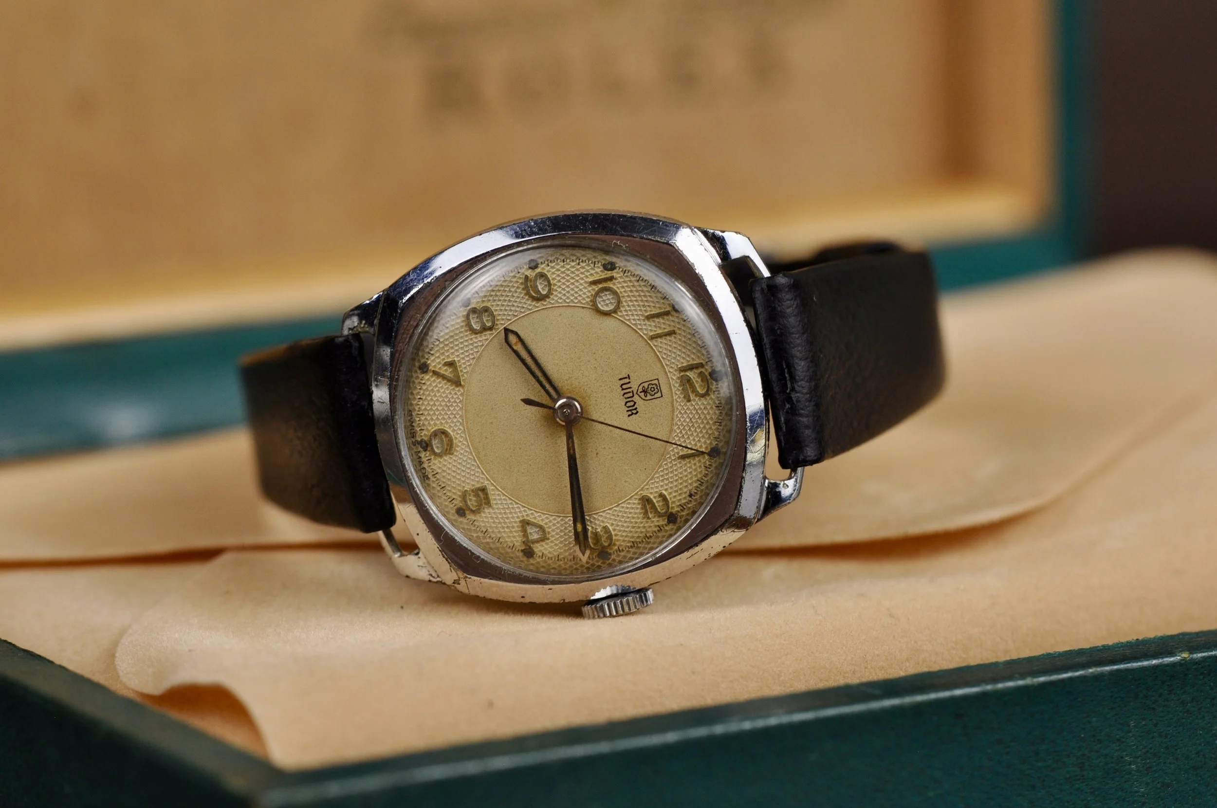 A vintage Tudor wristwatch with a beige dial, gold numerals, and black leather strap, resting on a tan cushion inside a green box.
