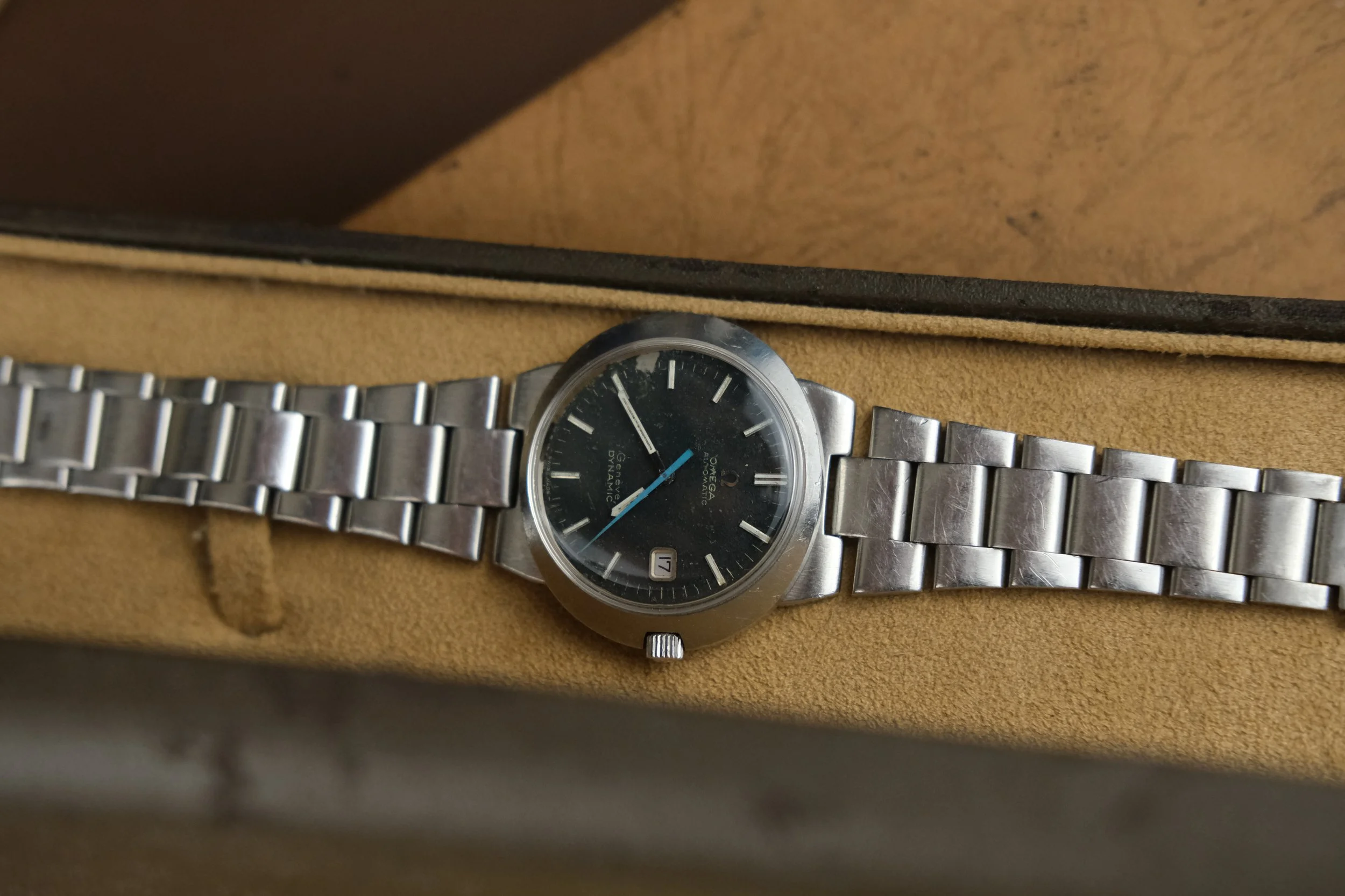 A vintage Omega Seamaster wristwatch with a silver band, black dial, and date window at 3 o'clock, resting inside a beige watch box.