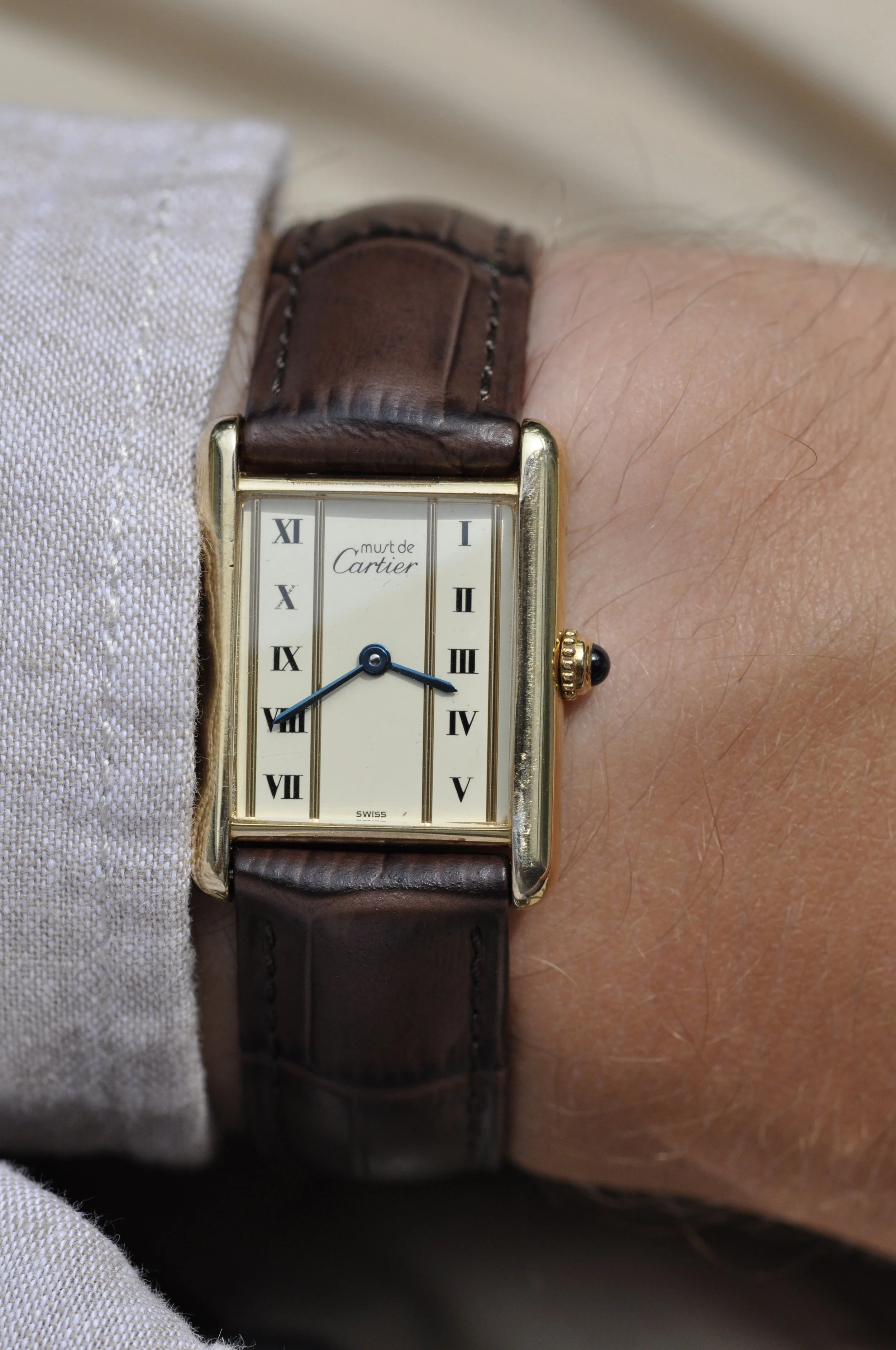 A person's wrist wearing a rectangular gold Cartier watch with a cream dial, blue hands, and a brown leather strap.