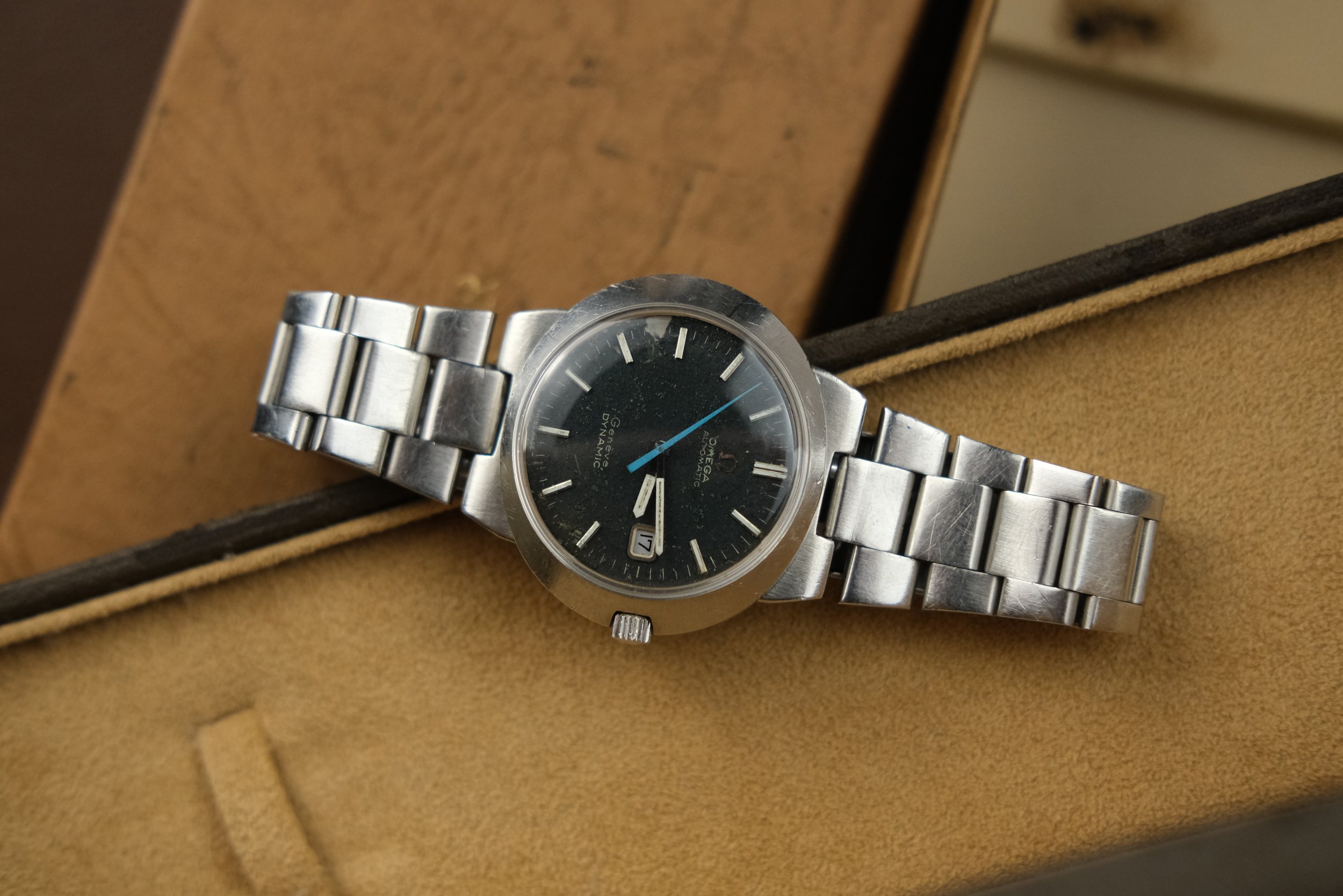 Vintage stainless steel Omega Genève Dynamic wristwatch with black dial, date window, and blue second hand, resting on a tan watch box.