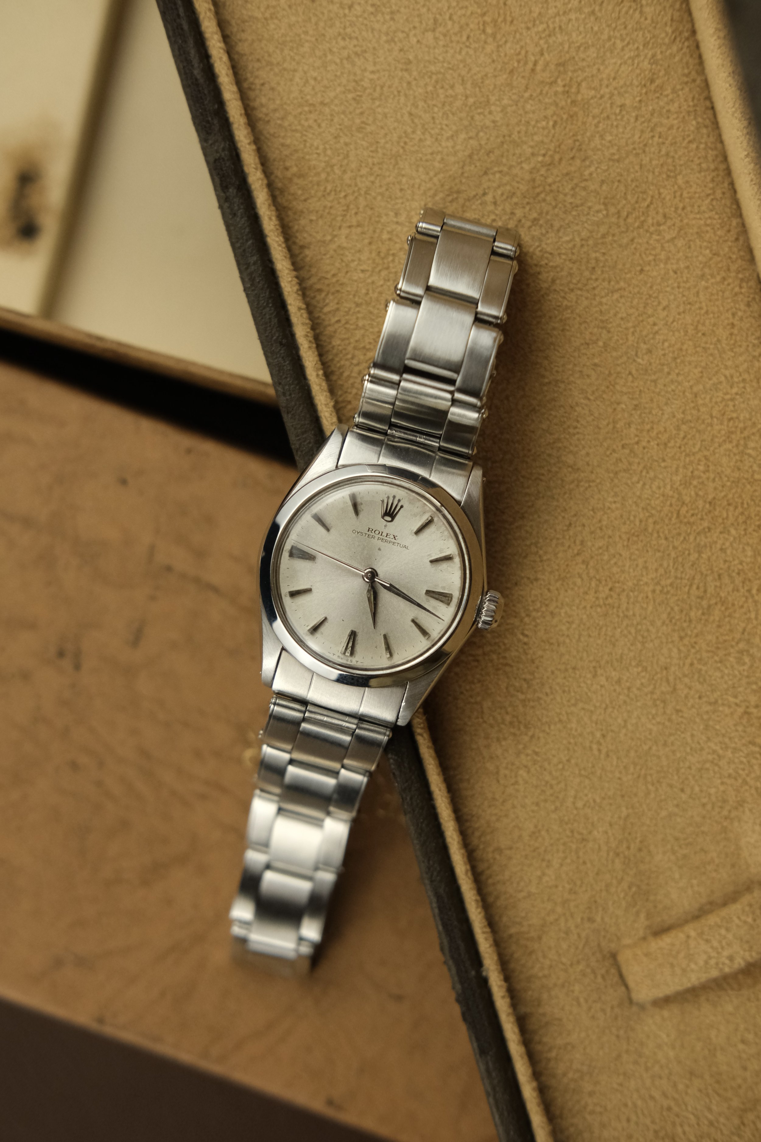 A silver Rolex Oyster Perpetual wristwatch with a metal band, placed on a beige cushioned surface inside a box.